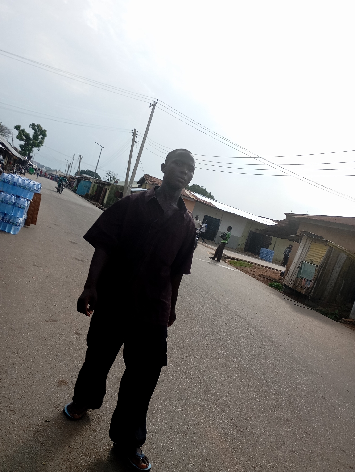 The image depicts a street scene in Shendam, Nigeria, under an overcast sky. In the foreground, a young Black male stands facing the camera, wearing a dark, short-sleeved collared shirt and dark trousers. He is wearing light blue and black flip-flops. To the left of the frame, a stack of blue-wrapped bottled water is visible. The street is paved and appears to be a commercial area, with low-rise buildings lining the right side of the road. Several other individuals are visible in the background, some walking and others near the buildings. A motorcycle is also present on the road. Utility poles with overhead wires run along the street. The environment appears to be a typical Nigerian urban or semi-urban setting.
