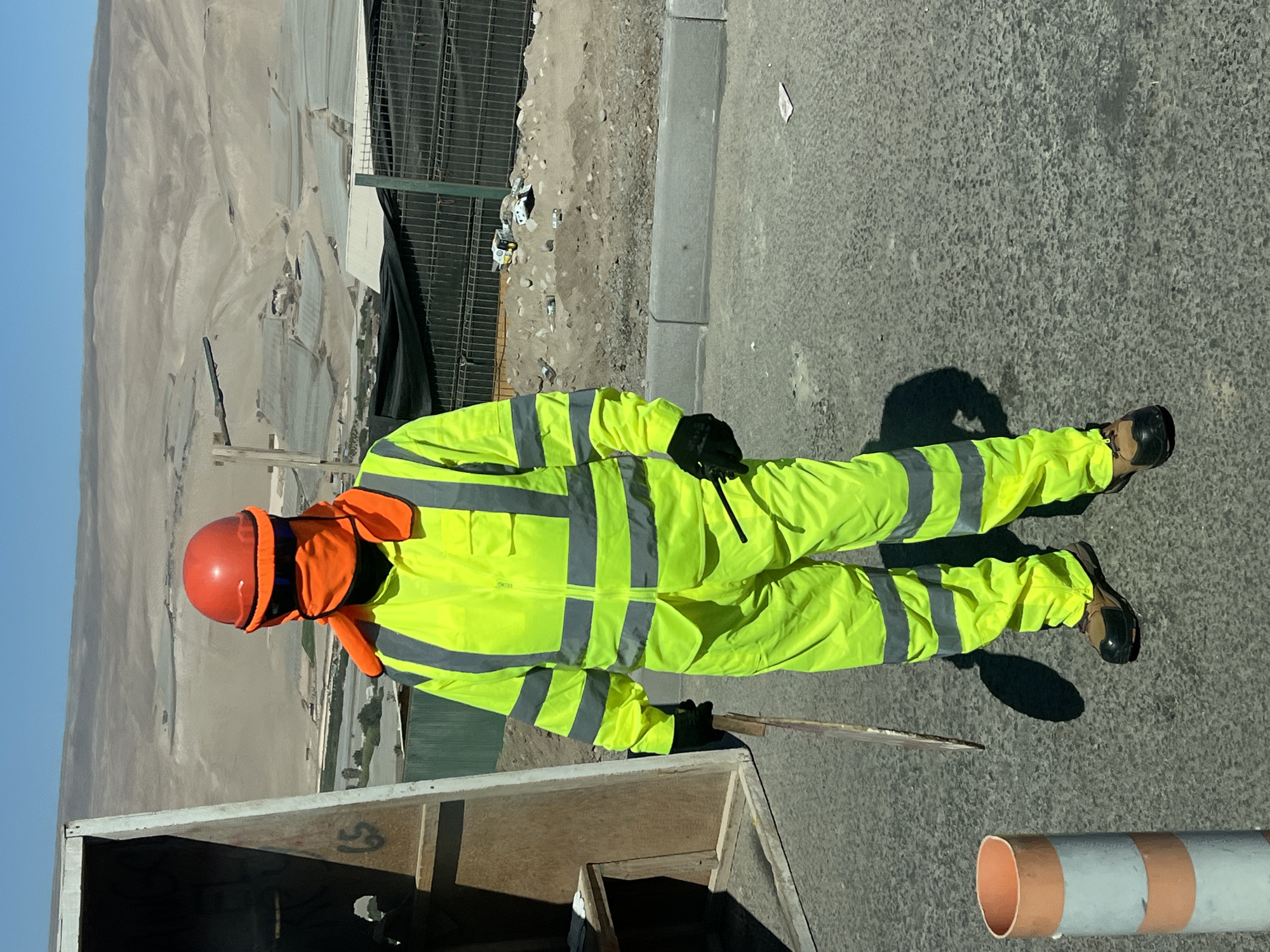 A person wearing a bright yellow safety suit, orange helmet, and gloves stands next to a structure on a paved surface. The person is also wearing an orange neck covering and dark-colored boots. In the background, a fenced area, a grey curb, and a desert landscape are visible under a clear blue sky. The location is Arica, Chile. A striped pipe is near the person's feet.