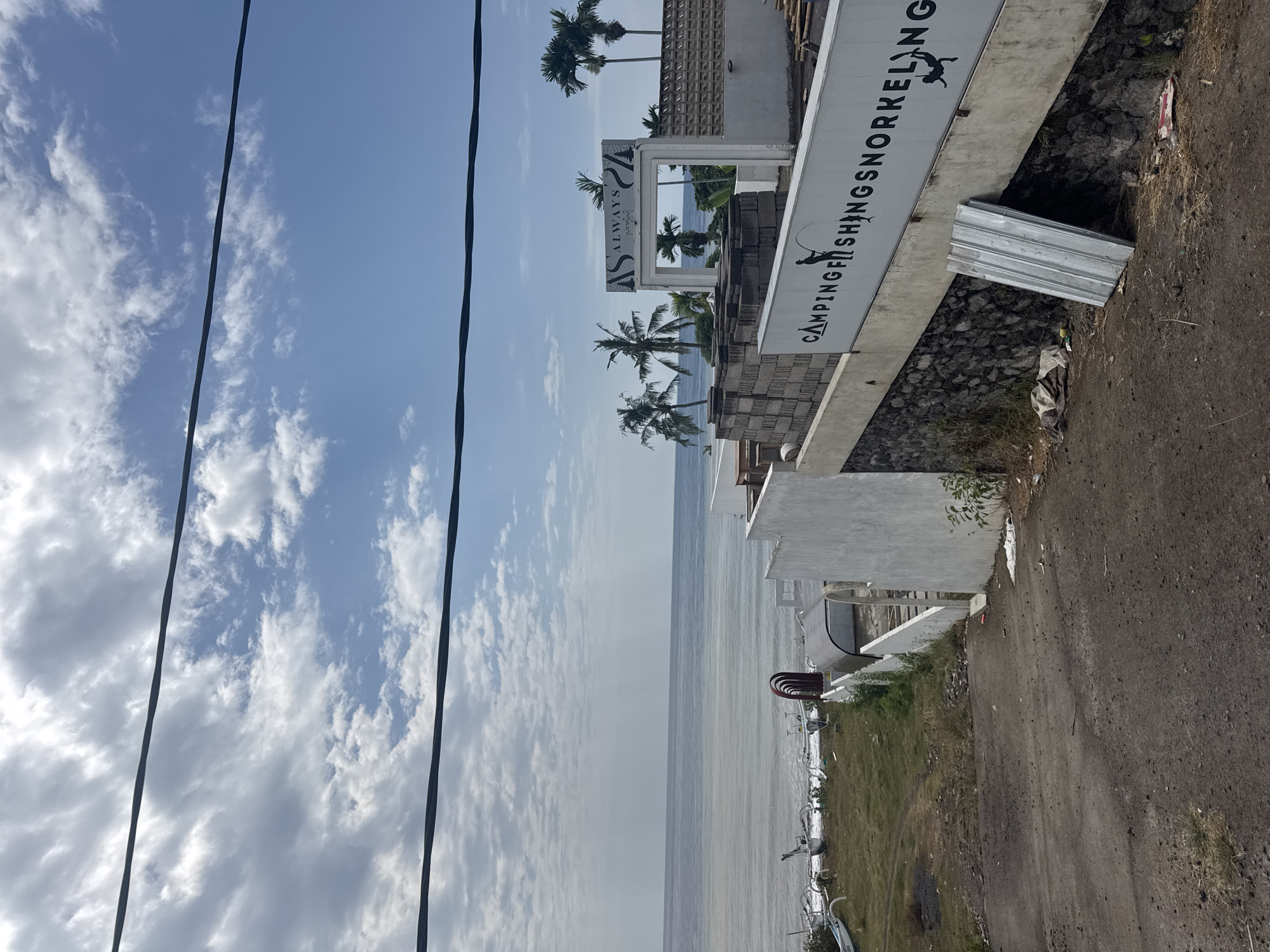 The image depicts a coastal scene in Abang, Indonesia, on a day with scattered clouds and blue skies. In the foreground, a dirt path leads down towards the ocean. To the right, a white wall with black lettering reads "CAMPING FISHING SNORKELING". Above this wall, a white structure with an arched opening displays a sign that says "ALWAYS". Palm trees are visible in the background, along with several traditional Indonesian fishing boats on the shore. The ocean stretches to the horizon. The scene appears to be a commercial establishment or resort area, possibly under development or in a state of disrepair given the discarded metal sheeting and debris on the ground.