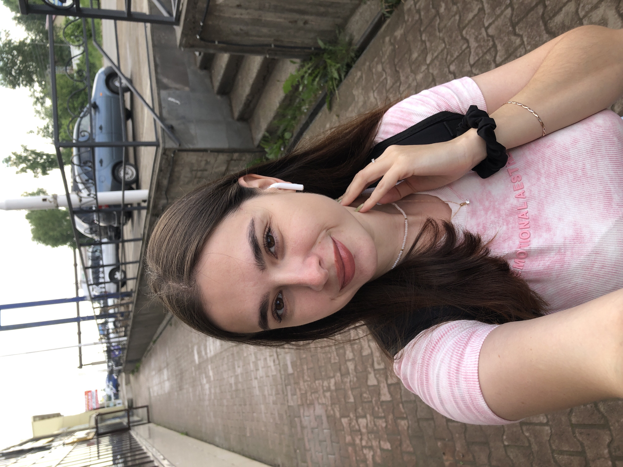 A young woman with long, dark brown hair and brown eyes takes a selfie outdoors. She is wearing a pink and white tie-dye t-shirt, a white beaded necklace, and a delicate gold bracelet. A white wireless earbud is in her right ear. She is standing on a brick sidewalk in front of a grey concrete wall with stairs and greenery. In the background, cars are parked and driving on a street. The sky is overcast. The location is likely Chaikovsky, Russia, given the context provided.