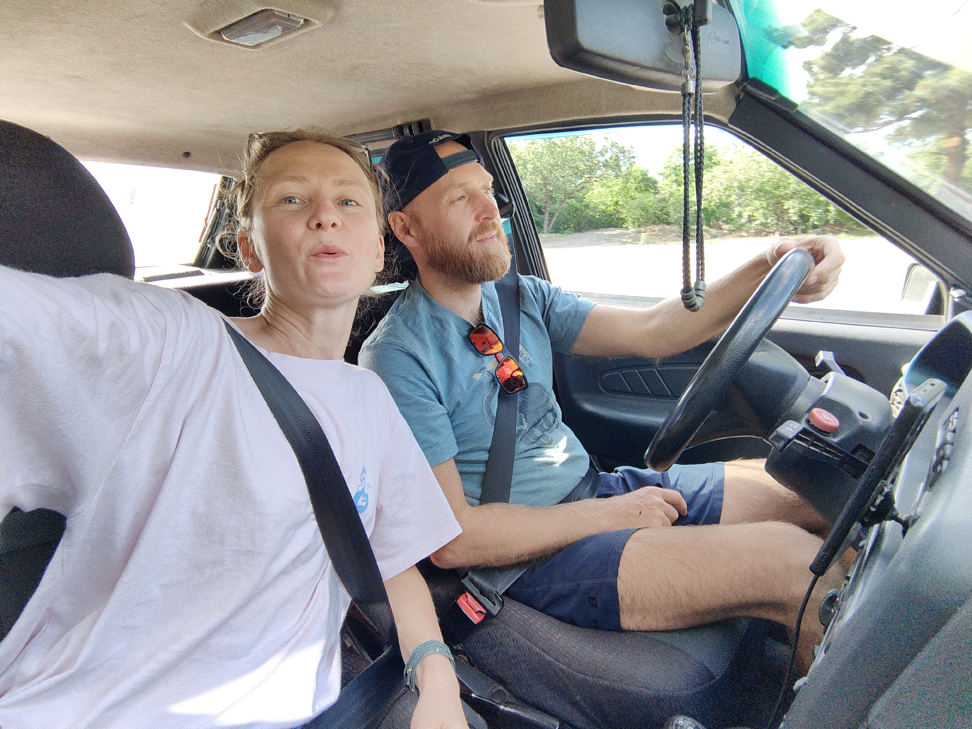 Two individuals, a woman and a man, are seated in the front seats of a car. The woman is in the passenger seat, facing the camera and making a kissing gesture. She is wearing a light pink t-shirt and a dark seatbelt. The man is in the driver's seat, wearing a blue t-shirt and dark shorts. He has a beard and is wearing a dark baseball cap. Sunglasses are hanging on his t-shirt. He is holding the steering wheel, indicating he is driving. The car's interior is visible, including the dashboard, steering wheel, and rearview mirror. Outside the car, trees and a road are visible through the windshield, suggesting the car is in motion. The scene appears to be outdoors, possibly on a road outside of a city, given the natural surroundings visible through the windows. Based on the provided context, the location is the city of Alushta, Ukraine.