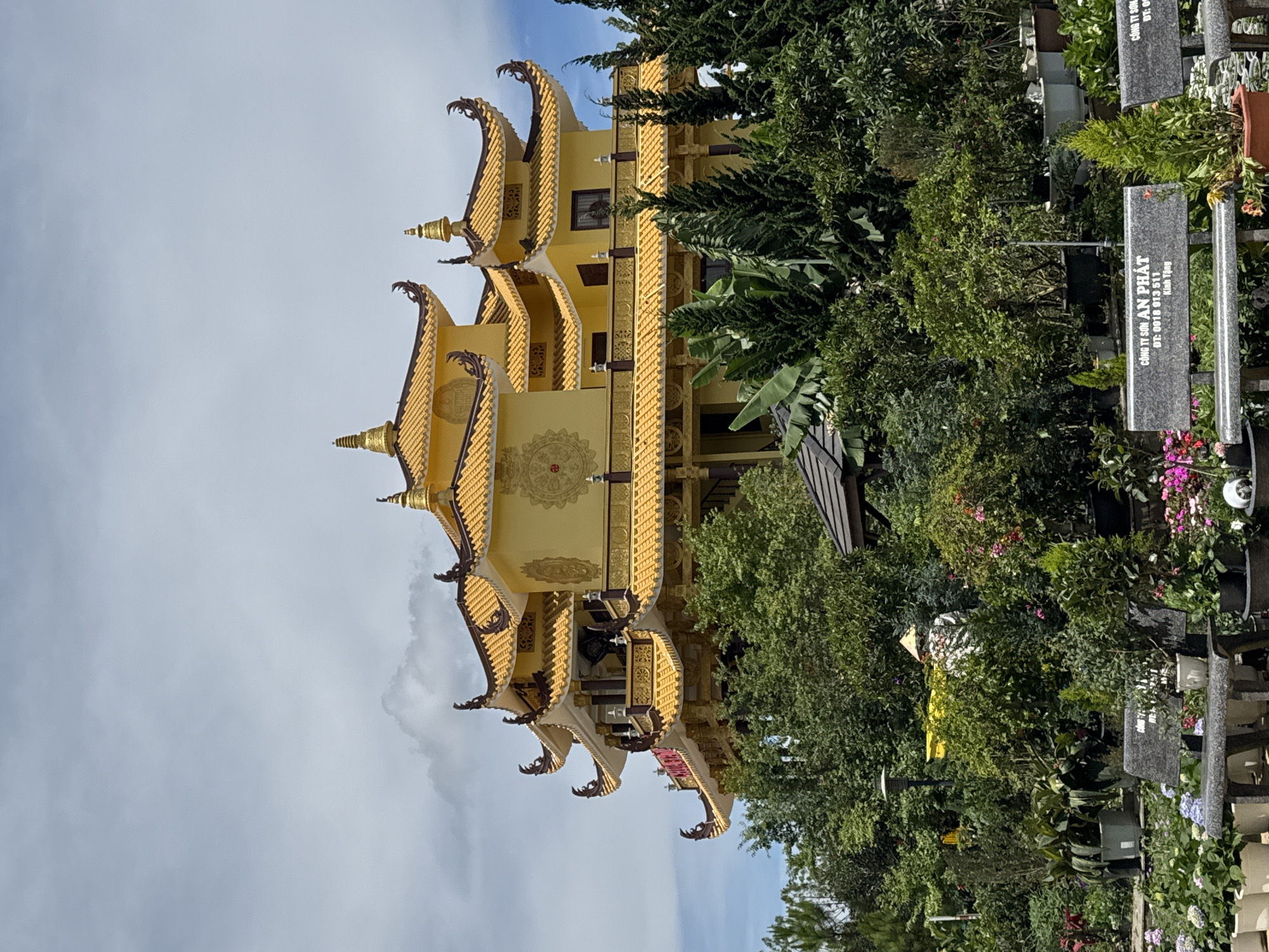 The image shows the Truc Lam Pagoda, a Buddhist temple in Da Lat, Vietnam. The pagoda, characterized by its yellow exterior and tiered, curved roof structure, rises prominently from the landscape. Lush vegetation, including a variety of trees and plants, partially obscures the view of the temple. In the foreground, grave markers are visible. A sky with clouds provides a backdrop to the temple.