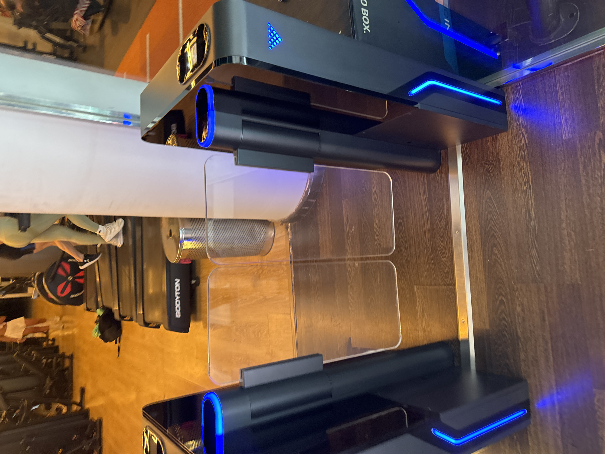 The image depicts an indoor gym environment, likely a fitness center in Murcia, Spain. In the foreground, there are two modern turnstile gates with blue LED lighting. The left gate is black and tall, with a curved top that glows blue. To its right, there's a clear plexiglass barrier. Further to the right, another turnstile gate, also black, features blue lighting and a triangular LED pattern. This gate appears to be part of a check-in or access control system, as a portion of text visible reads "BOX.".
In the mid-ground, a person wearing light-colored shorts and a light-colored top is visible walking away from the camera, towards the turnstiles. They are on a wooden floor, and behind them, a black treadmill is positioned. Another person is partially visible in the background, wearing green leggings and a black top, also walking on the wooden floor. To the left of the treadmill, a black backpack with a green item on top lies on the floor.
The environment appears to be a clean, well-lit gym with wooden flooring throughout. The background shows additional gym equipment, including weight machines and exercise stations, suggesting a functional fitness facility. The lighting is bright, with a focus on the illuminated turnstiles.
