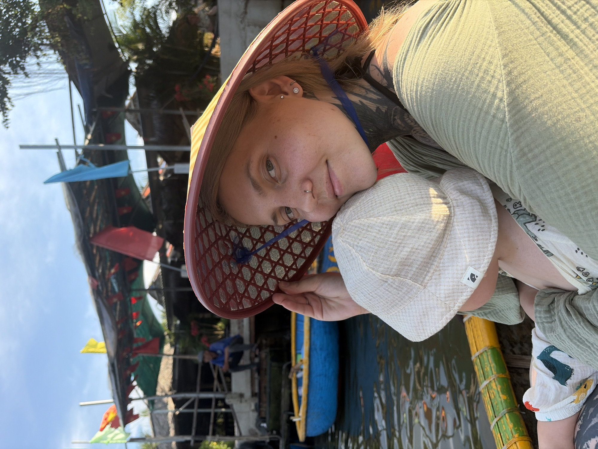 A light-haired woman and a small child are captured in a bright, outdoor setting, likely on or near a body of water. The woman, who has visible tattoos on her neck and shoulder, a nose piercing, and ear piercings, wears a light green ribbed top and a unique red mesh conical hat, akin to a traditional Vietnamese nón lá but with a modern design. She smiles softly, her head gently resting against the child's hat.
The child, mostly obscured, wears a light-colored hat with a small black tag featuring an abstract symbol and clothing adorned with colorful patterns, possibly dinosaurs.
The background reveals a body of water, likely a river or canal, with a blue boat partially visible. Along the opposite bank, structures, possibly houses or market stalls, are covered by canopies and decorated with small red, yellow, and blue flags. Green trees and foliage are visible against a clear, bright sky, indicating daytime. Another person in a blue shirt is seen sitting on a structure in the background. The scene suggests a relaxed atmosphere, typical of a waterway in Southeast Asia, consistent with the provided context of Đà Nẵng, Vietnam.