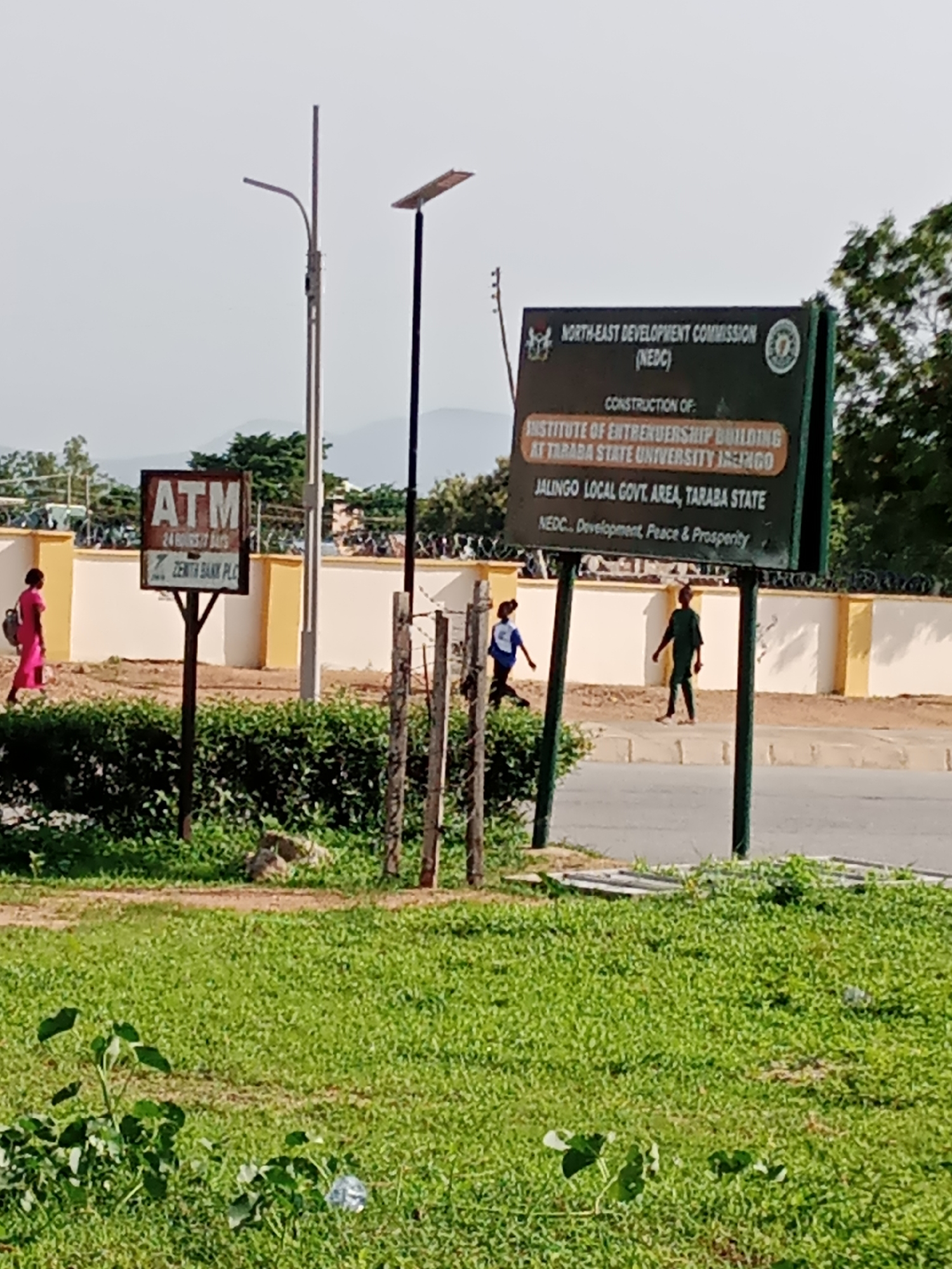 The image captures a street scene in Jalingo, Nigeria, likely in the vicinity of Taraba State University. In the foreground, a patch of green grass is visible, with some small plants and a discarded plastic bottle. Behind the grass, a road runs horizontally across the frame, with a sidewalk adjacent to it. Several individuals are present on the sidewalk. A woman in a pink dress walks from left to right, carrying a backpack. Further down the sidewalk, a young person in a dark green outfit walks from right to left, and another young person in a white and blue shirt and shorts runs from left to right.
Two signs are prominently displayed. On the left, a weathered sign reads "ATM" in large letters, with smaller text indicating "24 HOURS 7 DAYS" and "ZENITH BANK PLC". This sign is mounted on a metal pole. To the right, a larger, dark green sign with white lettering states: "NORTH-EAST DEVELOPMENT COMMISSION (NEDC)" at the top, followed by "CONSTRUCTION OF: INSTITUTE OF ENTREPRENEURSHIP BUILDING AT TARABA STATE UNIVERSITY JALINGO". Below this, it reads "JALINGO LOCAL GOVT AREA, TARABA STATE", and at the bottom, "NEDC... Development, Peace & Prosperity". This sign is also supported by metal poles.
The background features a cream-colored wall with yellow accents, topped with barbed wire fencing. Beyond the wall, trees and hills are visible under a bright, overcast sky. Two tall lamp posts, one with a solar panel on top, stand near the signs. The overall impression is of an outdoor public space in Jalingo, likely a campus or a public facility area, with ongoing construction indicated by the prominent sign.
