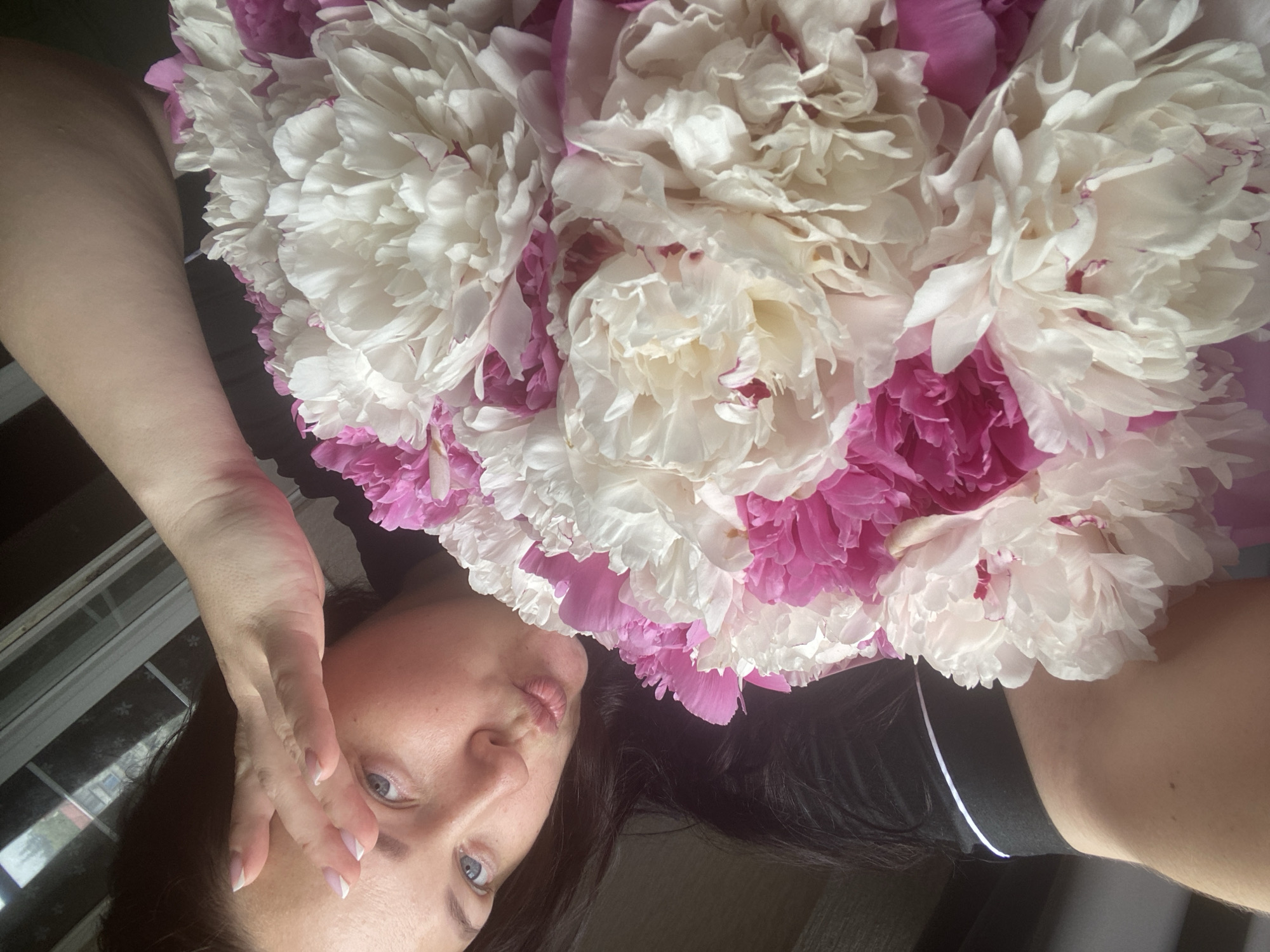 A selfie shows a woman with shoulder-length dark hair and blue eyes lying down. She is wearing a black shirt with a white stripe on the shoulder. Her left hand is raised near her face. She is positioned next to a large bouquet of white and pink peonies. The setting appears to be indoors, with a blurred view of windows and possible foliage visible in the background. There is no verifiable information to determine if the photo was taken in Sochi, Russia.