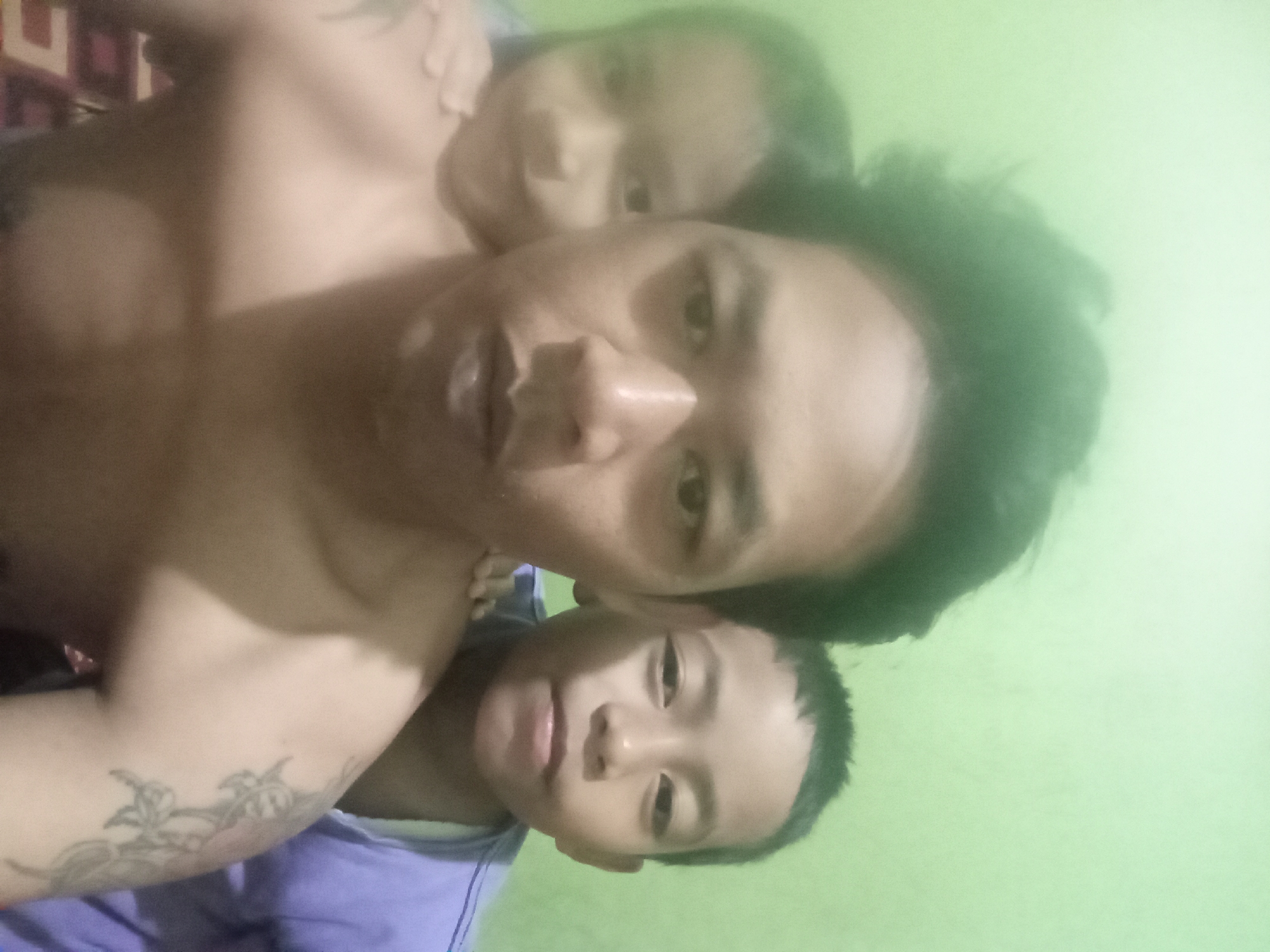 The media file is a low-resolution, candid photograph taken in Pagar Alam, Indonesia. The image features three individuals: an adult with a tattoo on their arm, and two children. The adult appears to be holding or embracing the children, with one child's face close to the adult's shoulder and another child positioned in front. The lighting suggests it might be daytime or indoor lighting, given the overall brightness and the light green wall in the background. The subjects are positioned close together, suggesting an intimate or familial moment. The overall tone of the image is casual and personal, likely a snapshot captured during a family gathering or a moment of affection. No specific objects or significant environmental details are clearly discernible beyond the basic elements of the people and the plain background. The image is too blurry to discern specific facial expressions or emotions with certainty. There is some visible text, "コ", in the top left of the image, which is likely a stylized character or logo.