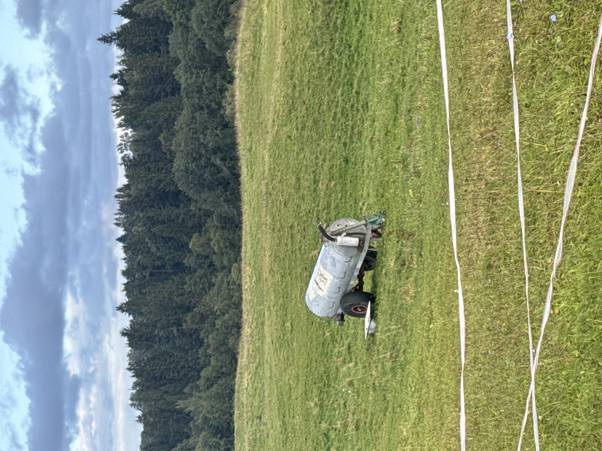 A metallic water trailer sits on a green grassy field. The trailer is cylindrical and mounted on two large wheels. In the foreground, a white electric fence runs horizontally across the image. In the background, a dense forest of dark green trees forms a backdrop against a cloudy sky. The scene appears to be located in or near Galinė, Lithuania, a rural area.