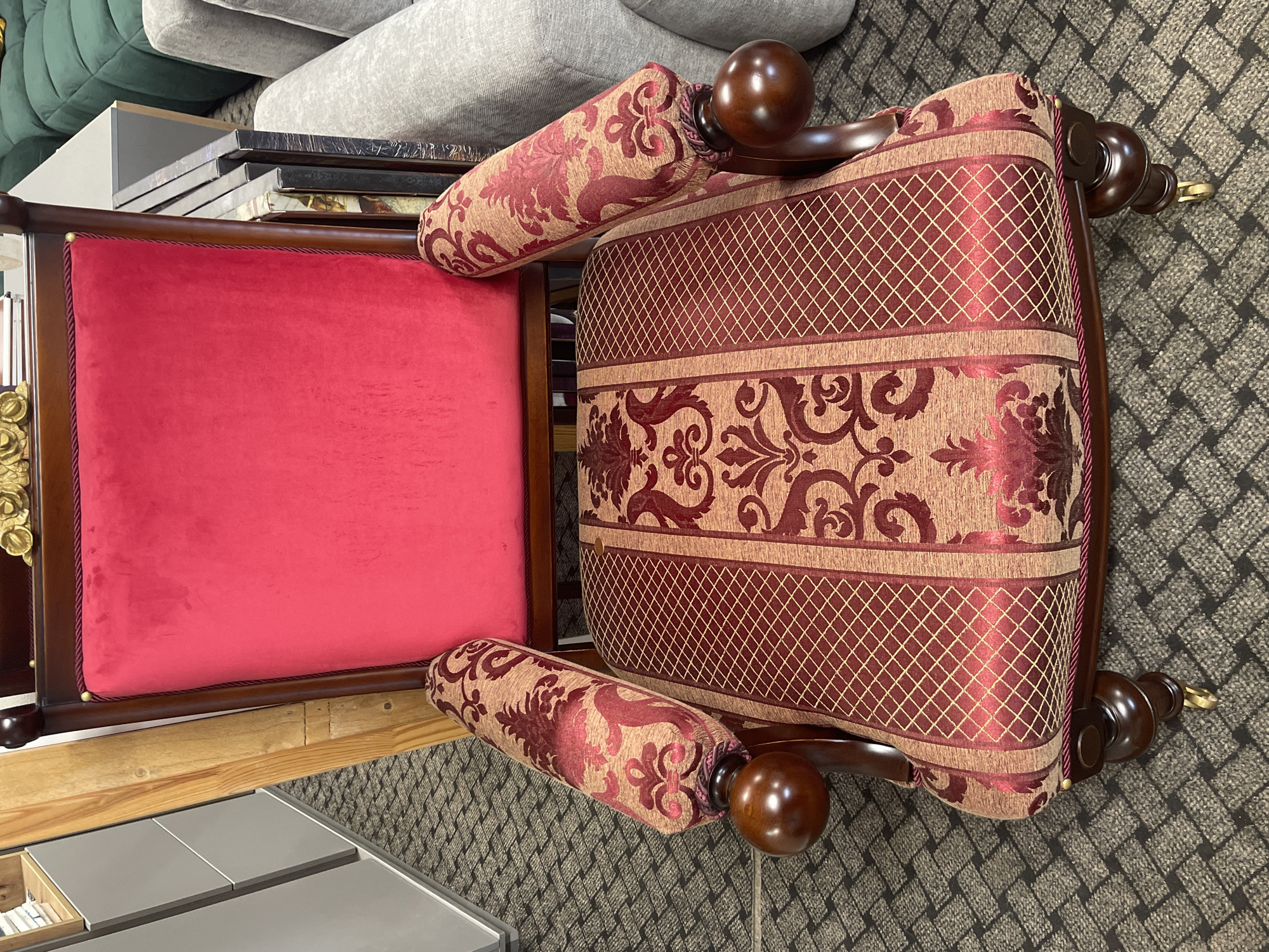 An ornate armchair with a vibrant red velvet backrest and intricately patterned upholstery on the seat and armrests is displayed in what appears to be a furniture store or showroom. The chair features dark wooden legs with casters and carved wooden armrests terminating in rounded finials. The seat and armrests are upholstered in a rich tapestry-like fabric with a maroon and gold damask pattern. The background shows various furniture items, including a green modular sofa and a grey sofa, along with shelving units. The floor is covered in a dark, patterned carpet. The lighting is bright, suggesting an indoor retail environment. There are no people present in the image.