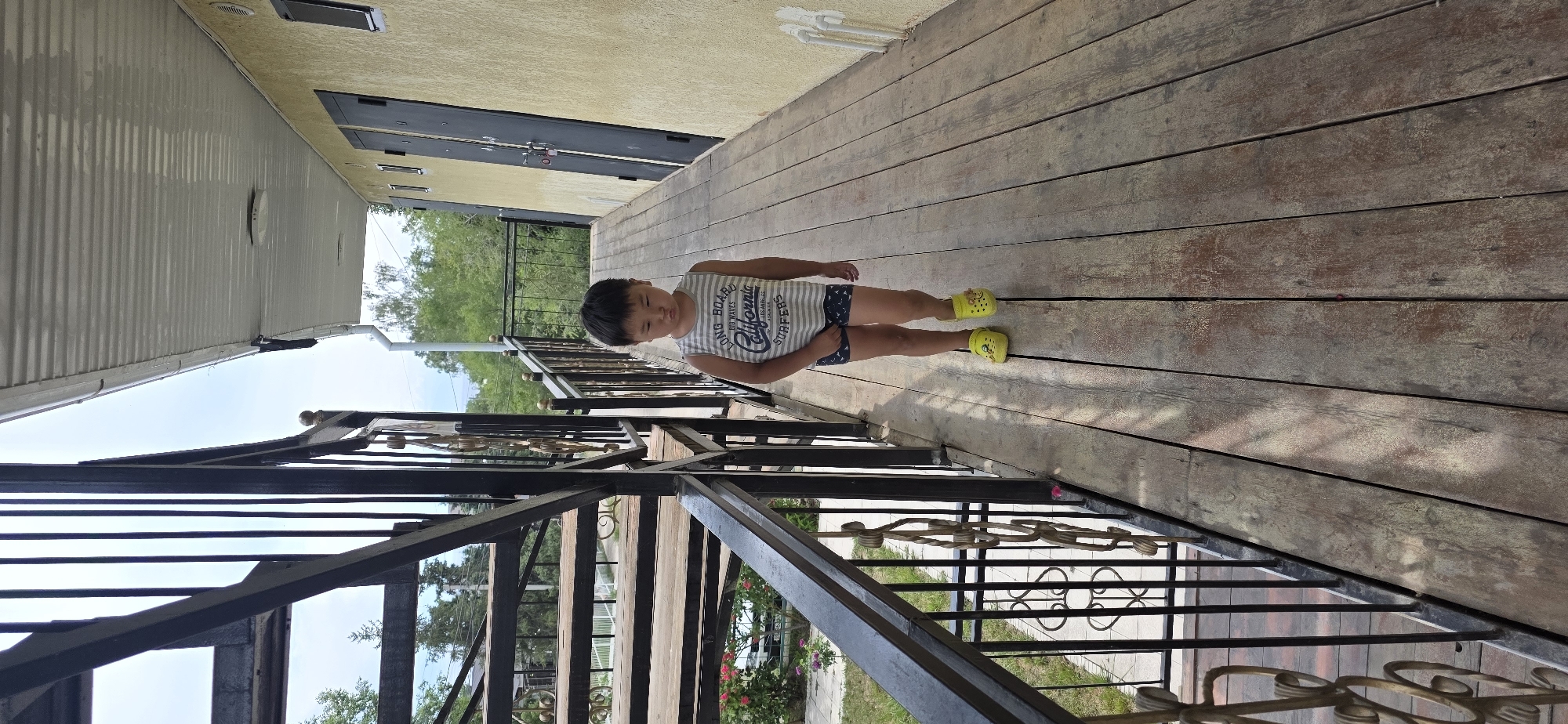 A young boy of East Asian descent, wearing a sleeveless striped t-shirt with the text "LONG BOARD BIG WAVES California SURFERS" and patterned shorts, stands on a wooden walkway. He is also wearing yellow Croc-style shoes. The boy is facing the camera with his lips pouted. The walkway appears to be an exterior balcony or porch, with a black metal railing to the left and a yellow building with dark doors to the right. Behind the railing, a set of wooden stairs is visible, with trees and greenery in the background. The scene is set in Бактуу-Долоноту, Kyrgyzstan, under a bright sky.