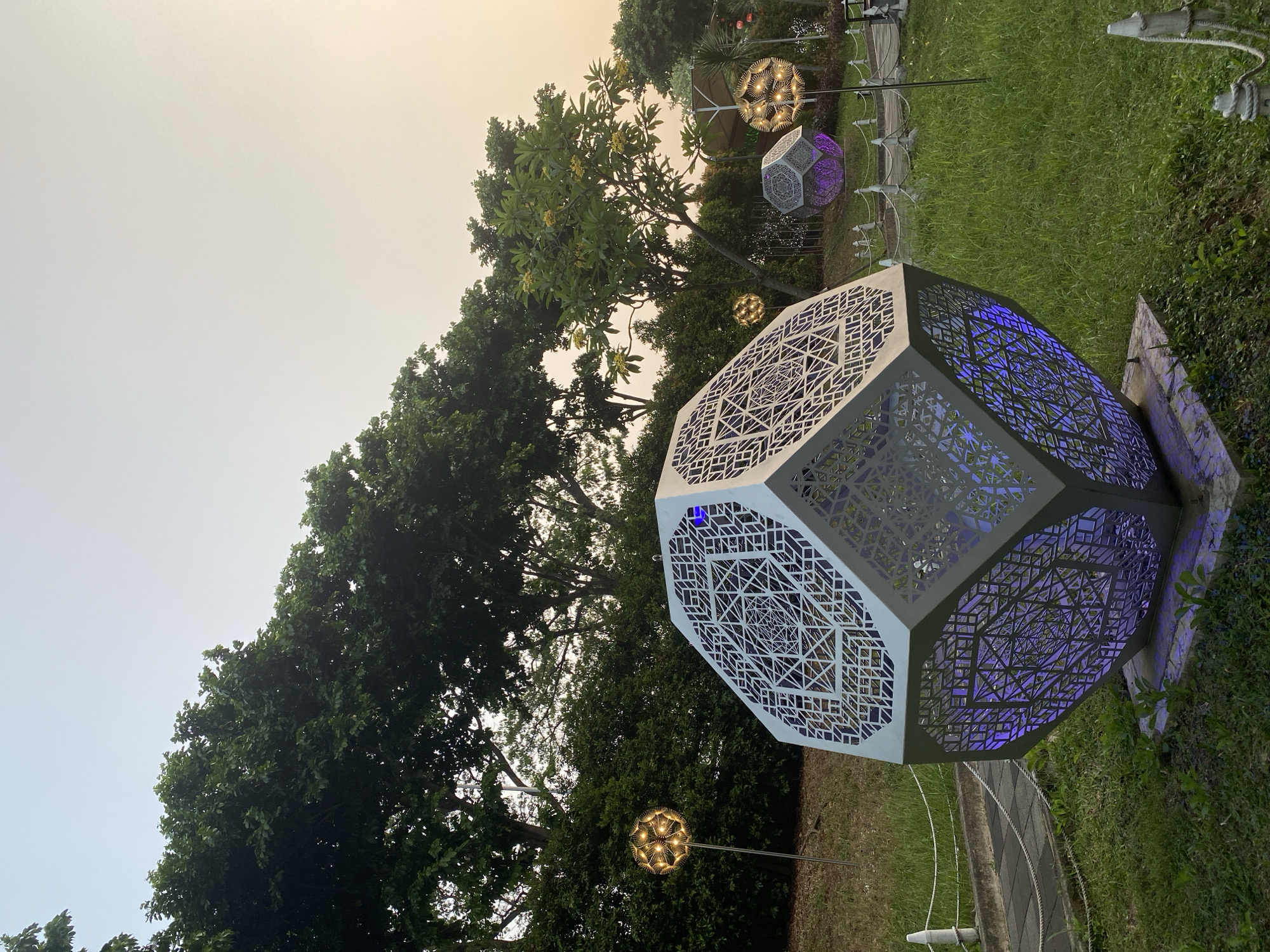 The image depicts an outdoor park scene at dusk, featuring several illuminated geometric sculptures. In the foreground, a large, white, faceted sculpture with intricate cutouts is prominently displayed. It is illuminated from within with a vibrant purple light. The sculpture rests on a concrete base.
Behind the main sculpture, other similar geometric forms are visible, one of which also emits a purple glow. To the left and right of the foreground sculpture, spherical, wireframe lanterns with warm, yellow lights on poles are scattered throughout the park. These lanterns have a dandelion-like appearance.
The environment is a grassy park area with mature trees and dense green bushes lining the background. A paved walkway with white rope barriers separates the grassy areas. The sky is a soft, muted orange and yellow, indicating either sunrise or sunset. The overall atmosphere suggests an art installation or a festive light display in a public space. The location is identified as East Jakarta, Indonesia, specifically within the Taman Mini Indonesia Indah park, in the area surrounding the Keong Mas building.