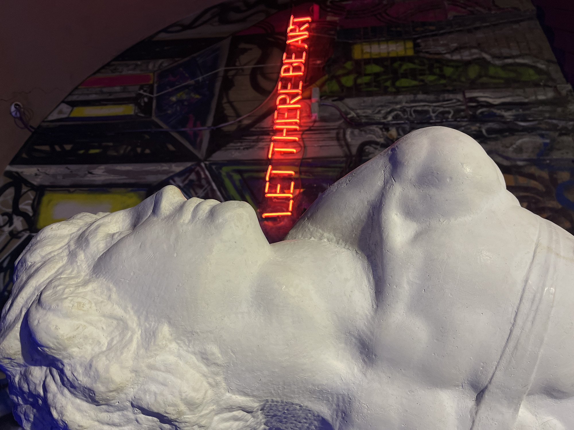 The image presents a close-up view of a white, textured sculpture of a reclining male figure, positioned in front of a wall adorned with graffiti or mural art. A neon sign reading "LET THERE BE ART" vertically aligns behind the sculpture, creating a stark contrast between the classic form and modern art expression. The setting might be an art gallery or public space within Saint Petersburg, Russia, considering its vibrant art scene and cultural emphasis. The foreground sculpture dominates the frame, while the background's mural adds context. The lighting emphasizes the contours of the sculpture and the brilliance of the neon sign.