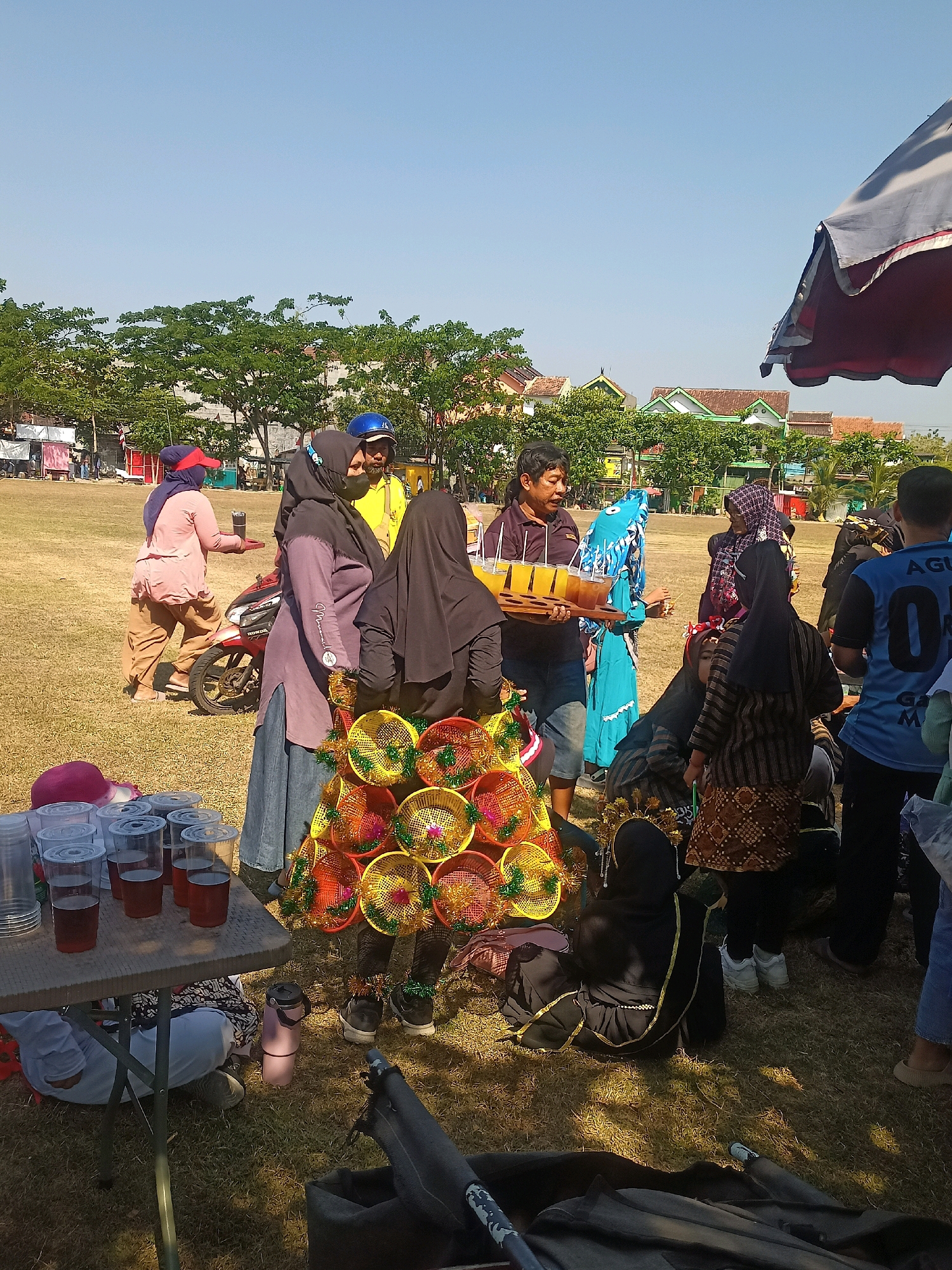 An outdoor, daytime, medium shot captures a lively scene on a grassy field in Sumberlawang, Indonesia. A group of people, some wearing Islamic headscarves, are gathered around a table laden with drinks in plastic cups and a tray of beverages. In the foreground, a colorful, conical structure adorned with woven baskets and tinsel stands prominently. Several individuals are engaged in conversations or observing the surroundings. Behind them, a red motorcycle is parked, and further in the background, trees and buildings suggest a semi-rural or suburban setting. The sky is clear and blue.
