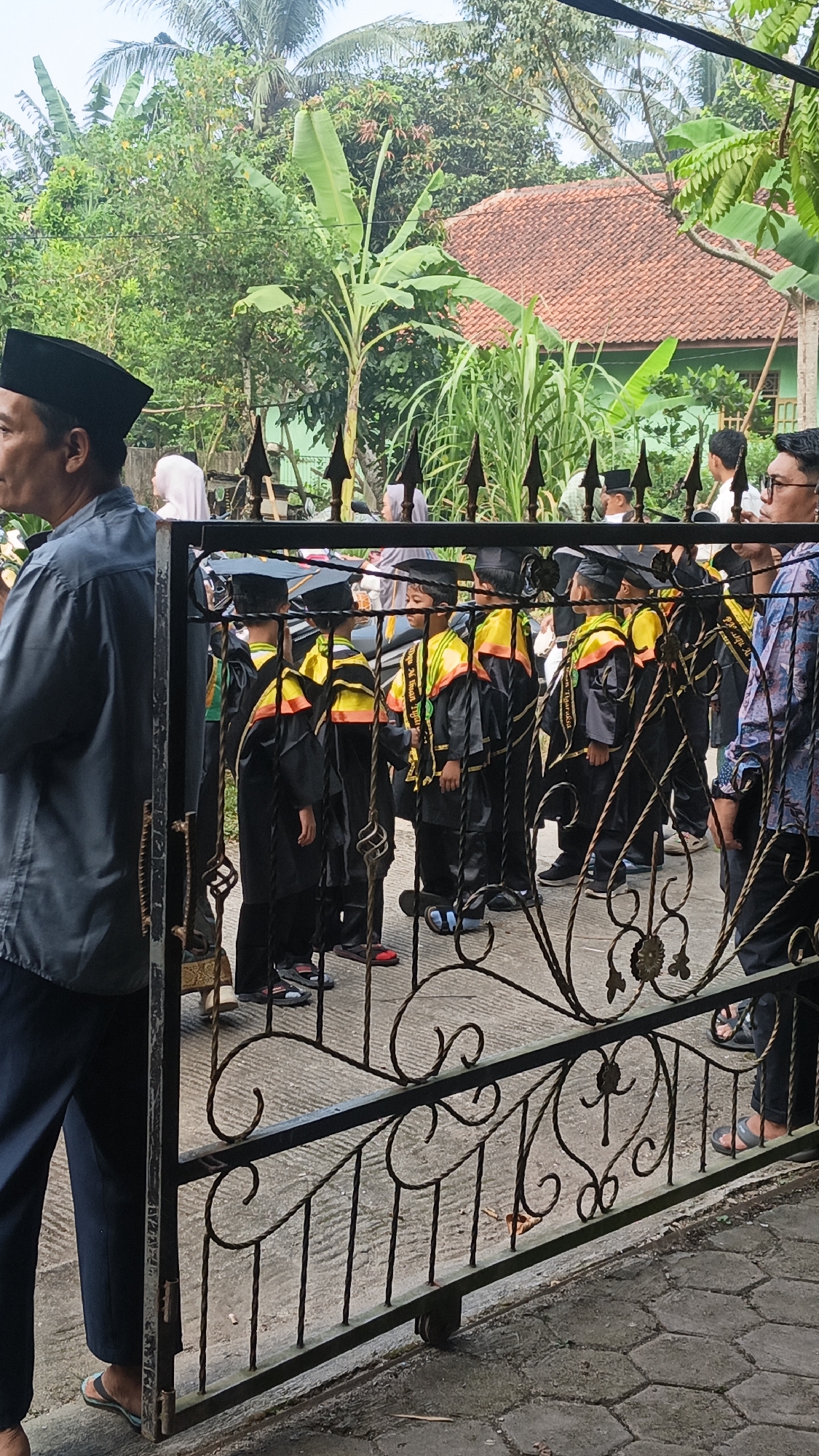 An exterior medium shot captures a group of children dressed in black graduation gowns and caps, standing behind a black wrought-iron gate. The children are lined up, facing the viewer's left, with several adults visible behind and to the right of them. The children are wearing black gowns with yellow and orange sashes draped over their shoulders. They appear to be waiting or observing something. In the background, lush green trees and a building with a terracotta-tiled roof are visible, suggesting a residential or school setting. The ground is paved with hexagonal tiles. The scene suggests a graduation ceremony or a related event taking place in Tigaraksa, Indonesia.