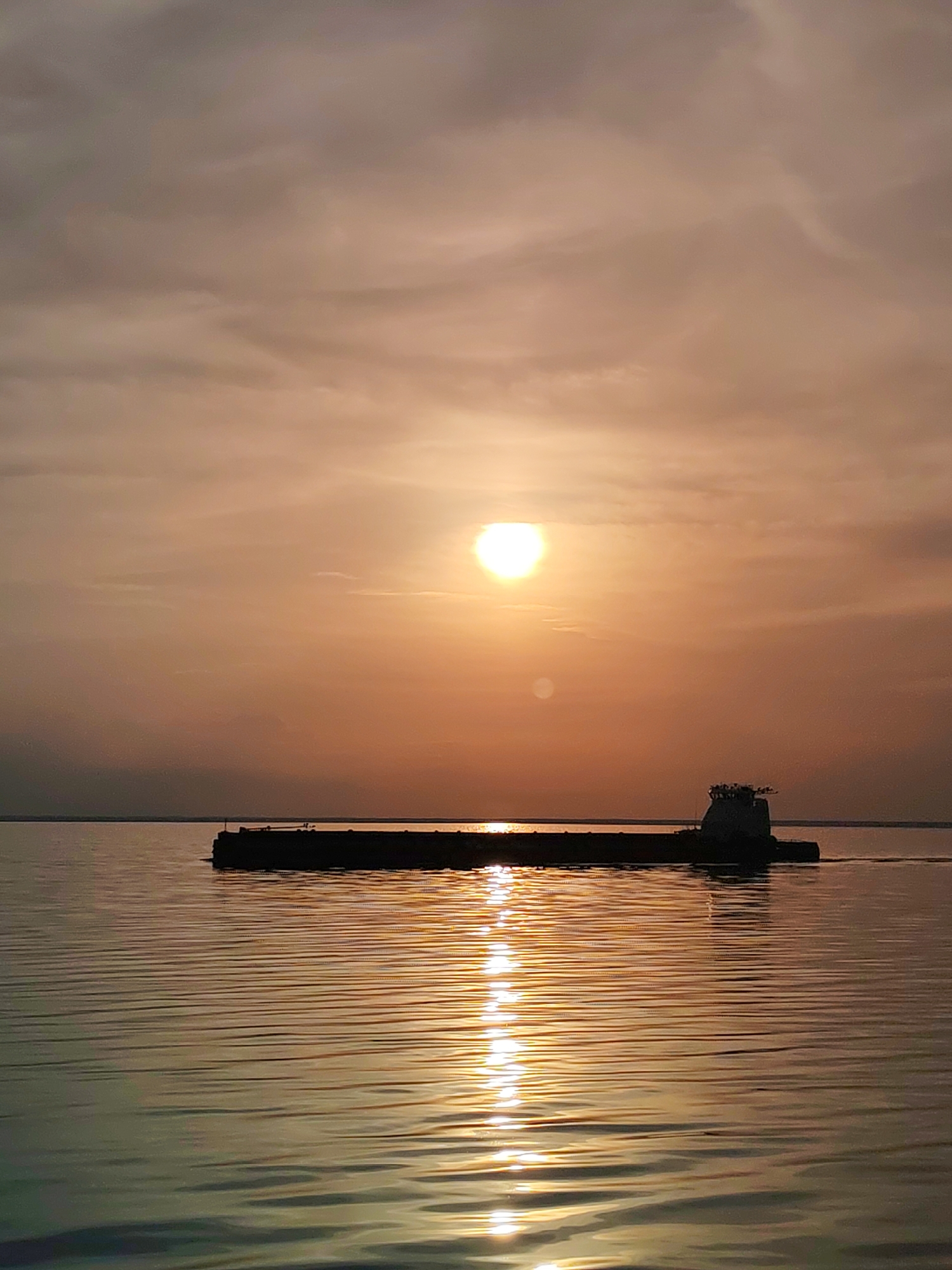 A barge is sailing on a body of water with the sun reflecting in the water. The sky is a hazy orange color, with clouds visible. The sun is a bright circle in the center of the sky. The barge is dark and silhouetted against the bright background. The water is calm, with small waves reflecting the sunlight. It is near Belozersk, Russia.