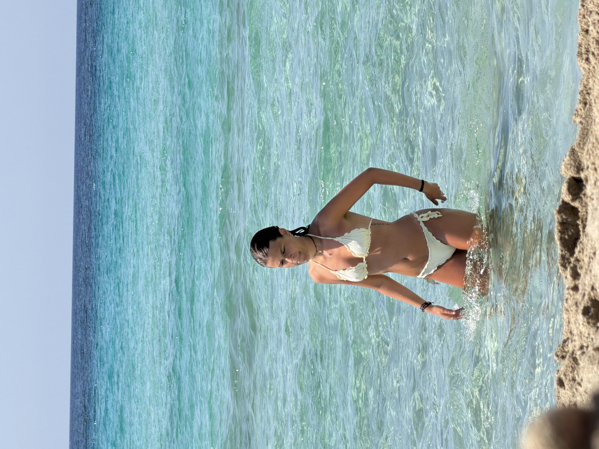 A young woman in a white bikini is standing in shallow, clear turquoise water at a beach. The water is calm with gentle ripples. The background shows the ocean transitioning from a lighter turquoise to a deep blue, with a clear sky above. The foreground features a sandy beach. The woman is looking slightly downwards and to her left.
