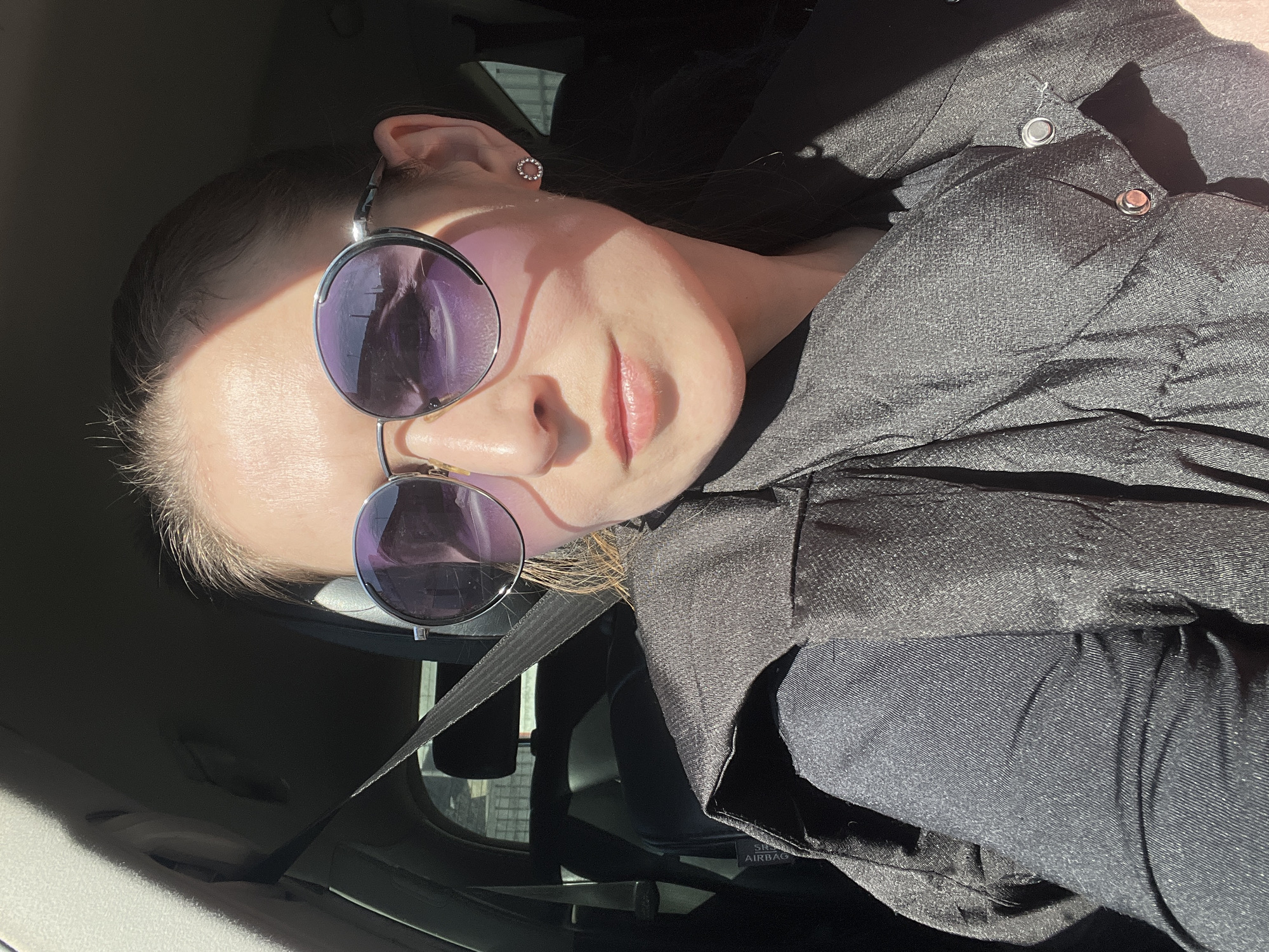 A woman is depicted from the upper torso, seated in what appears to be a car interior, likely in Votkinsk, Russia. She is wearing round, purple-tinted sunglasses that cover her eyes, and a small, circular, sparkly earring in her left ear. Her lips are slightly parted. She is dressed in a dark, possibly black, textured jacket or vest over a dark top. A seatbelt is visible across her chest and shoulder.
The image is captured in bright, direct sunlight, creating strong highlights on her skin and clothing, particularly on her forehead, cheek, and right shoulder, while other parts of the car interior and her person are in shadow. The reflection in her sunglasses shows outlines of what could be buildings or trees, consistent with an outdoor setting. The interior of the car is dark, with visible details including a headrest and parts of the dashboard. Text partially visible on a car component, likely a seat, reads "AIRBAG". The woman's expression is neutral.