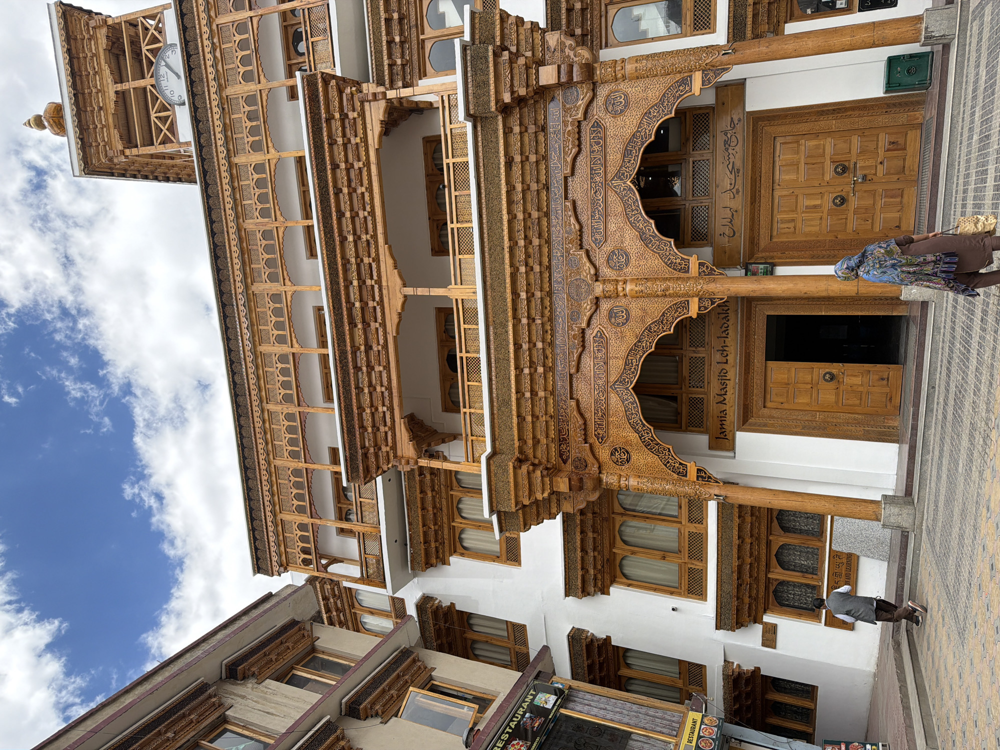 The image shows the Jamia Masjid mosque in Leh, India, on a cloudy day. The mosque features intricate wooden carvings on its facade and a clock tower. Two people are visible: one man walking away from the camera and a woman facing the mosque's entrance. The street in front of the mosque is paved with patterned tiles. Signs for a restaurant are visible on the left side of the image.
