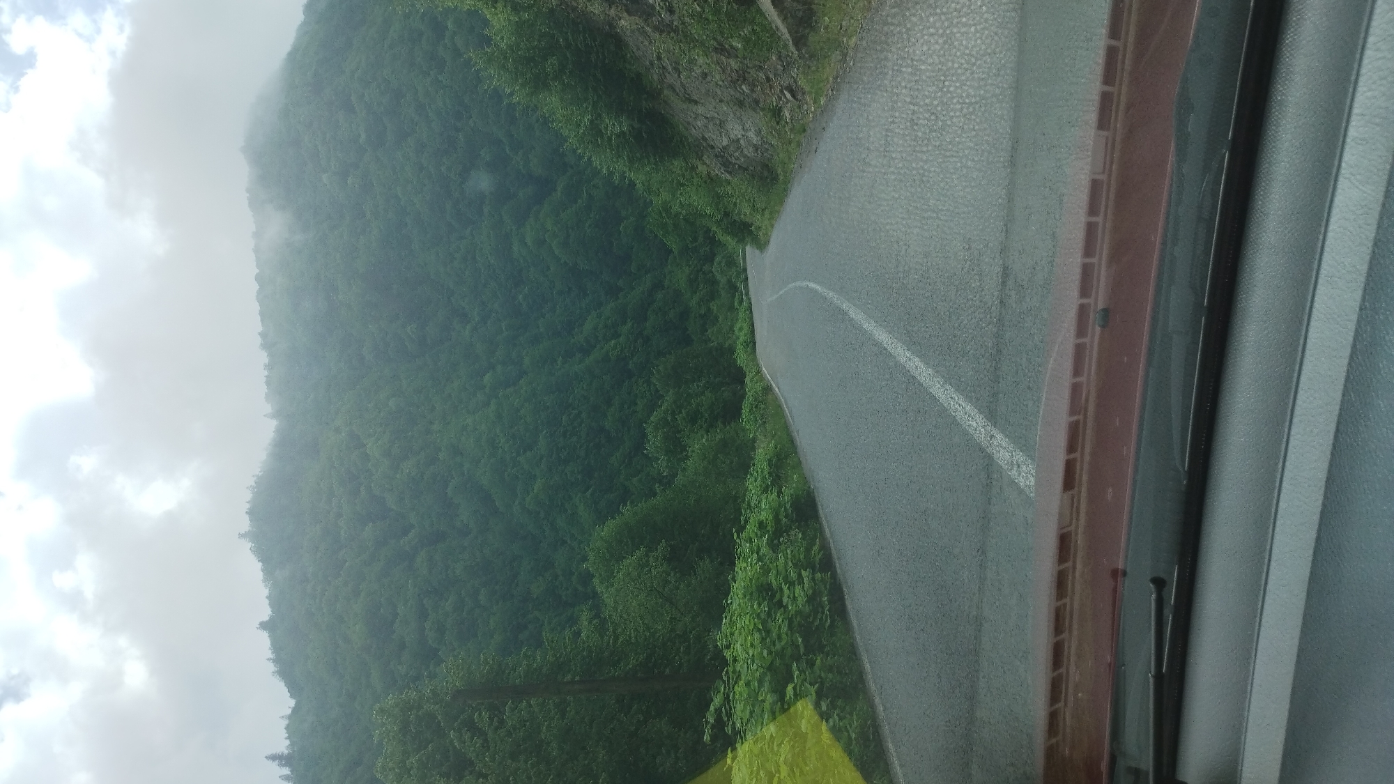 The image shows a view from inside a vehicle, likely a car, looking out onto a paved road. The road curves to the left. A white dividing line runs down the center. To the left of the road is a densely forested mountainside shrouded in mist or low-hanging clouds. The sky is overcast with grey clouds. Parts of the dashboard and windshield of the vehicle are visible in the foreground. Location information (Kostylivka, Ukraine) is provided, but not visually verifiable within the image.
