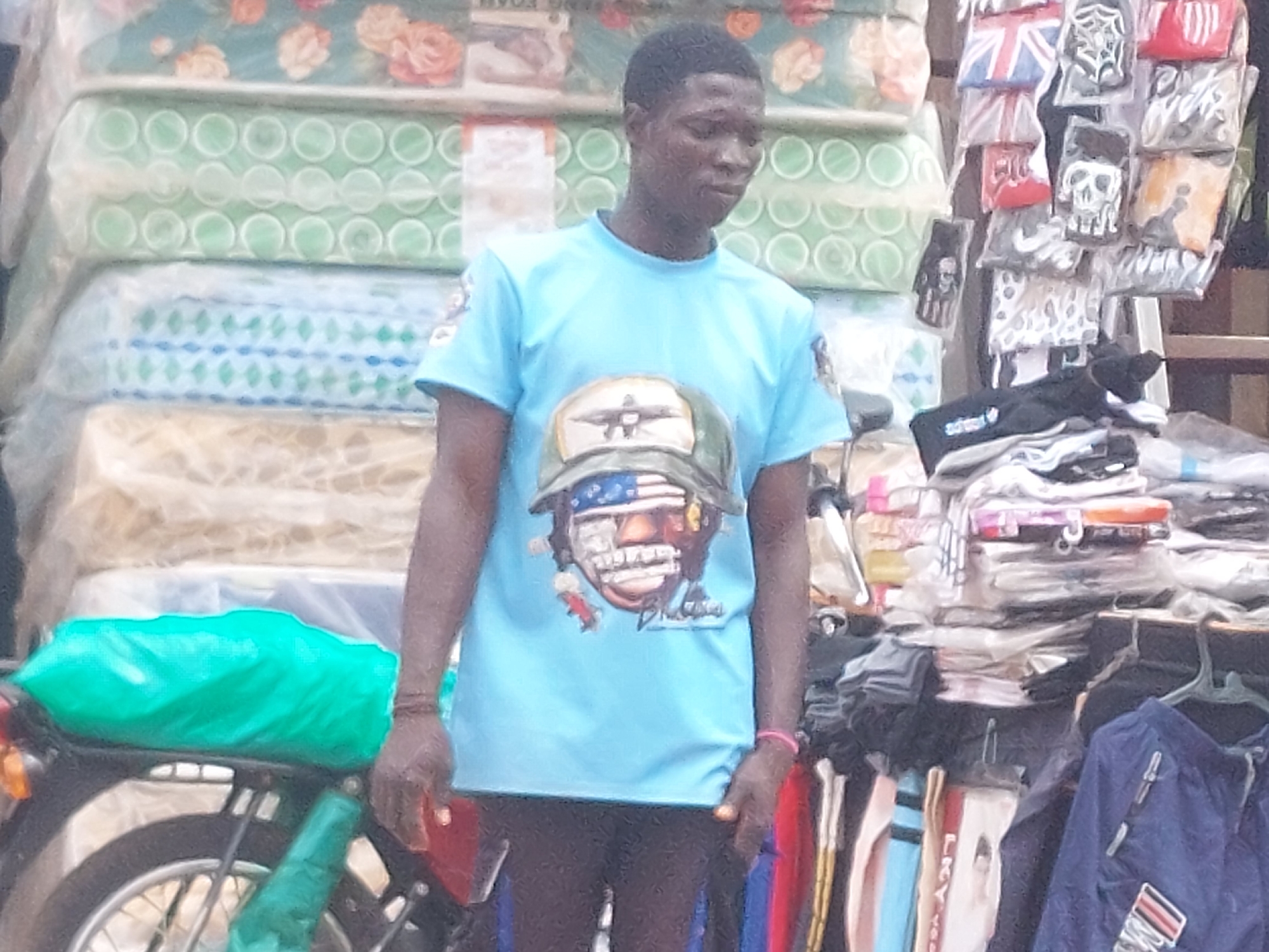 This image captures a male individual standing in what appears to be an outdoor market setting in Unknown, Unknown.
The central figure is an adult male with a dark complexion and short, dark hair. He is wearing a light blue short-sleeved t-shirt featuring a graphic print of a military-style helmet with an American flag design on the face shield and a dark green helmet shell. An emblem, possibly an aircraft, is visible on the helmet, and the word "Pilot" is faintly discernible below the graphic. He wears dark trousers or shorts and a thin pink bracelet on his right wrist (viewer's right). His left hand (viewer's left) holds a red rectangular object, possibly a mobile phone. He is looking downwards and slightly to his right.
To the left of the individual, several mattresses or foam pads are stacked vertically, each encased in clear plastic. The uppermost visible mattress features a floral pattern with orange roses on a teal background. Below it are two mattresses with a repeating light green circular pattern, followed by one with a blue and green diamond pattern. The lowest visible mattress has a light, textured pattern.
To the right of the individual, a display rack showcases various hanging items, likely accessories or small garments, mostly packaged in clear plastic. Designs include a Union Jack flag, a white skull on a black background, a spiderweb on a black background, and a red item possibly bearing an Adidas logo. Below these hanging items, stacks of folded merchandise are visible. Further down, a dark blue garment, possibly a jacket or track pants, with a red and white striped patch is seen on a hanger. A black item, possibly a bag or glove, with "adidas" printed in white, rests on a lower surface.
In the bottom left foreground, a section of a bicycle or motorcycle is visible, including a wheel and frame. A bright green plastic cover is draped over the seat or rear rack, and another green object is near the wheel.
The scene is illuminated by natural light, indicative of an outdoor or semi-outdoor commercial area.