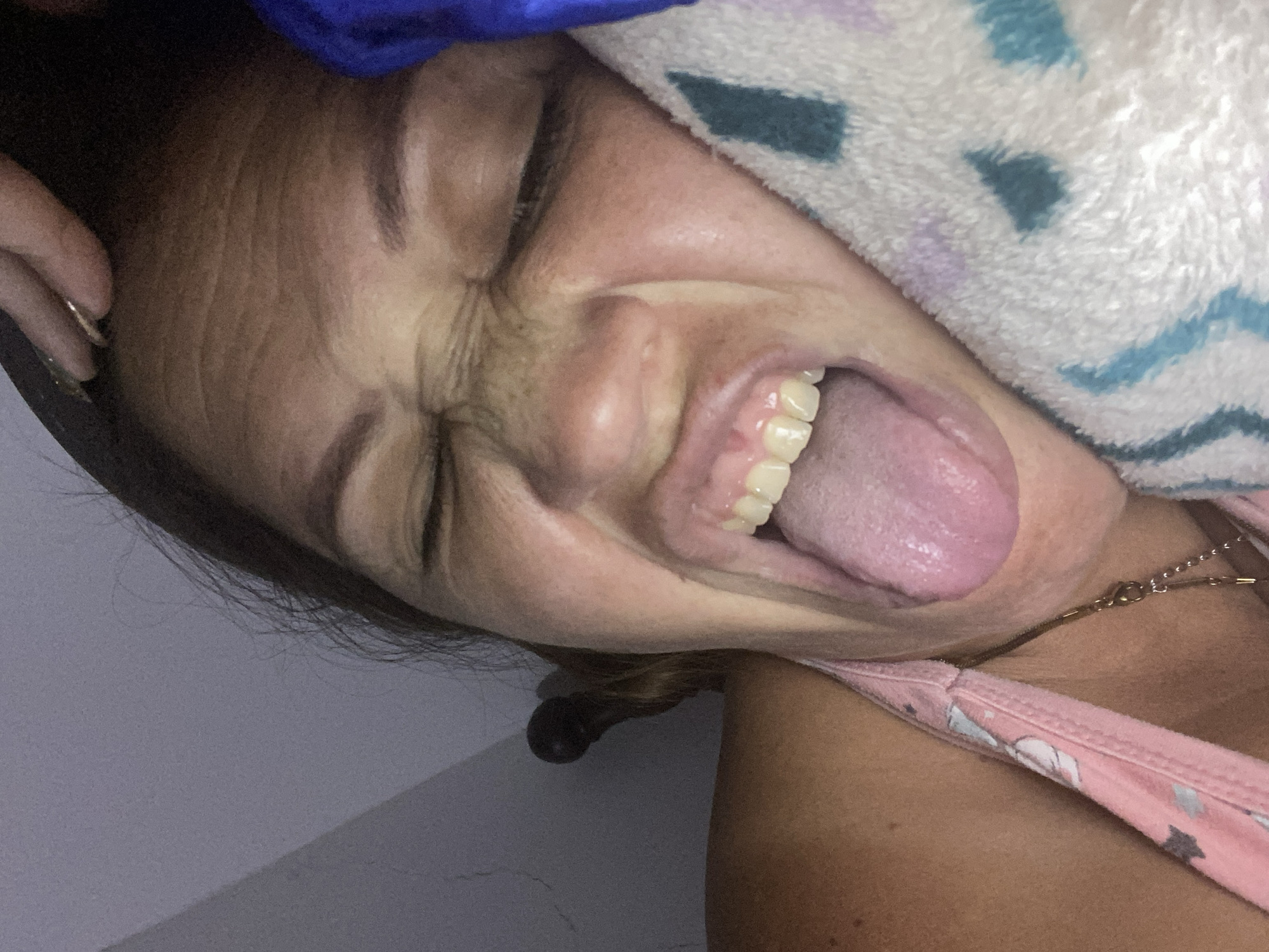 The media file is a close-up, low-angle shot of a person's face, with their eyes squeezed shut and their mouth open, sticking out their tongue. The subject appears to be under a blanket or towel with a blue and white patterned design. The person's face shows wrinkled skin around the eyes and nose, suggesting a strong expression. Their teeth are visible and appear to be somewhat yellow. A gold necklace is visible around their neck, and they are wearing a pink top with a floral or star pattern. The lighting is somewhat dim, suggesting it could be indoors or nighttime. The background is a plain, light-colored wall. The overall impression is one of playfulness or silliness, given the facial expression and the way the tongue is protruding. No text is visible in the media file.