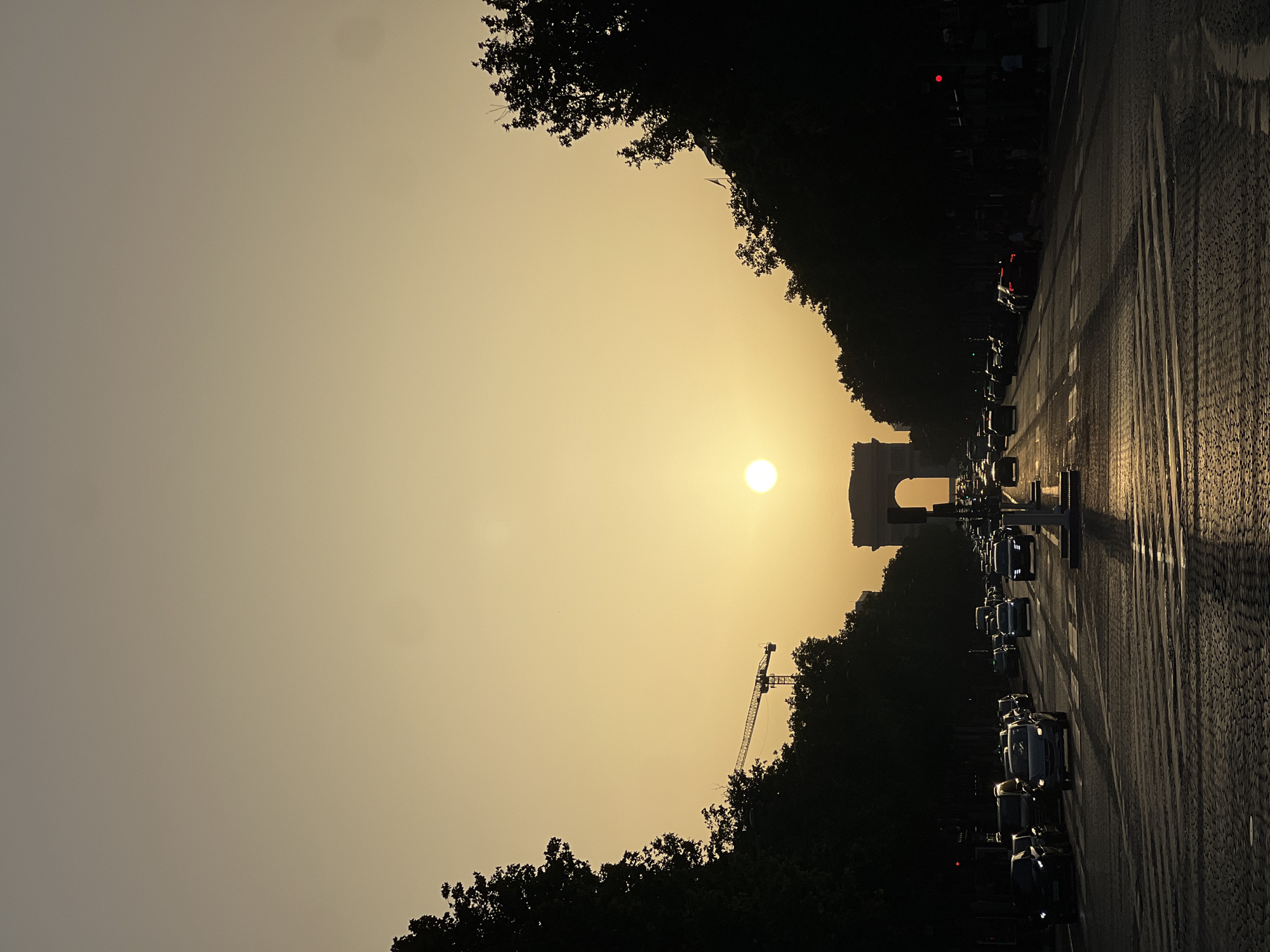 The image captures a street scene in Paris, France, at sunset. The Arc de Triomphe is visible in the distance, centered on the street. The sun is positioned in the sky, creating a strong glare and backlighting the scene. Cars are lined up on the street, indicating traffic. Trees line both sides of the street, silhouetted against the sky. A crane is visible among the trees on the left side of the image. The street itself appears to be paved with cobblestones. The sky is a yellow-orange hue, reflecting the sunset.