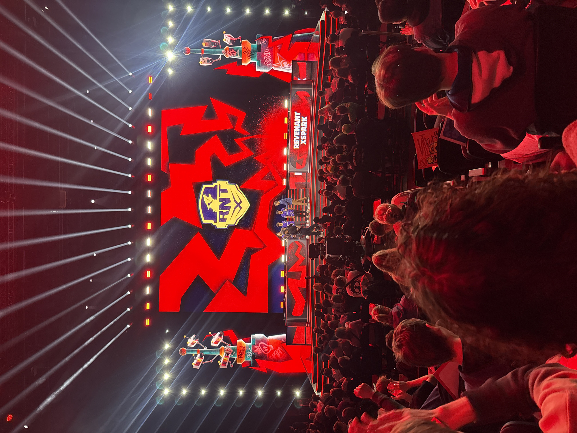 The image shows an indoor event, possibly a gaming tournament, with a large audience. A massive screen displays the logo "RNT" in the center, flanked by red lightning bolt-like designs. Above the central stage area, the words "REVENANT XSPARK" are visible. The screen is illuminated with bright spotlights creating beams of light that shoot upwards. The audience is dense, with people of varying ages visible. Some individuals appear to be holding up objects or signage, though details are obscured. The overall lighting is dark, with the stage and screen being the primary sources of illumination. The image was taken at an event in Berlin, Germany.