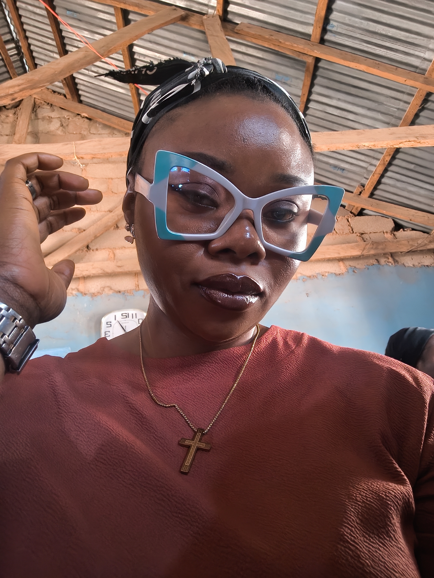 A close-up, high-angle shot depicts a woman in Nabatum, Nigeria. She wears large, turquoise and white framed glasses, a headscarf, a rust-colored top, and a gold cross necklace. A silver watch is visible on her left wrist. A wall clock showing approximately 5:57 is mounted on a blue and white wall behind her. The background includes a roof structure of corrugated metal and exposed wooden beams.