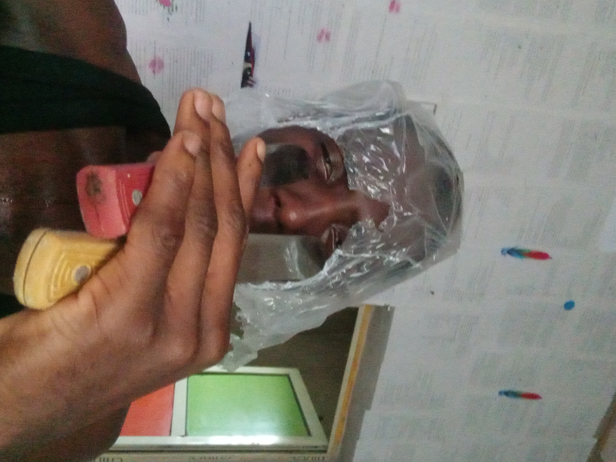 A person holds a handheld tool with a red and yellow handle in their left hand, positioned near their body. The person's face is visible inside a clear plastic bag. The background is a patterned wall with what appears to be a window frame below. The location is Akwete, Nigeria.