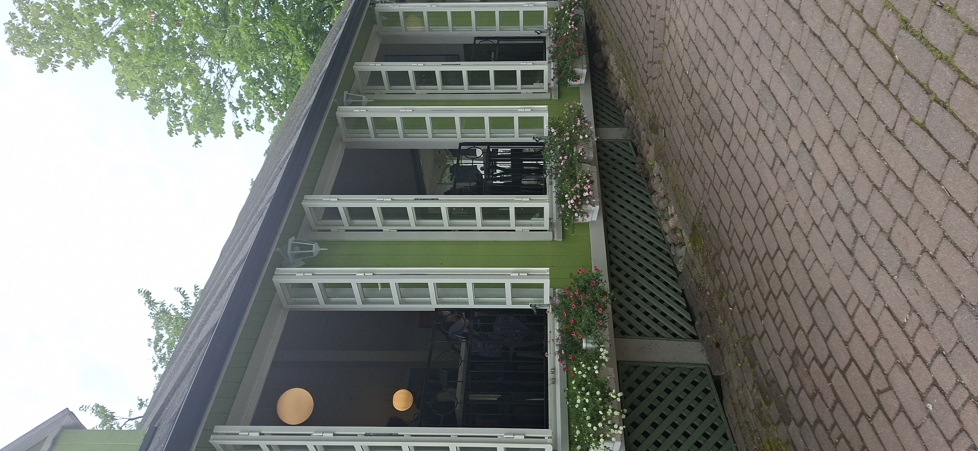 This is a photograph of a green building with large, open windows, possibly a cafe or restaurant, located in Naantali, Finland. The building has a dark-colored roof and is adorned with flower boxes containing pink and white flowers. The area in front of the building is paved with reddish-brown brickwork. Inside the building, through the open windows, tables and chairs are visible, suggesting an interior seating area. Two round, yellow pendant lights hang inside the building. Trees with green foliage are visible behind and to the side of the building, indicating an outdoor, possibly park-like setting. The sky is overcast and light-colored. A green lattice structure is visible below the flower boxes, covering the lower part of the building's exterior.