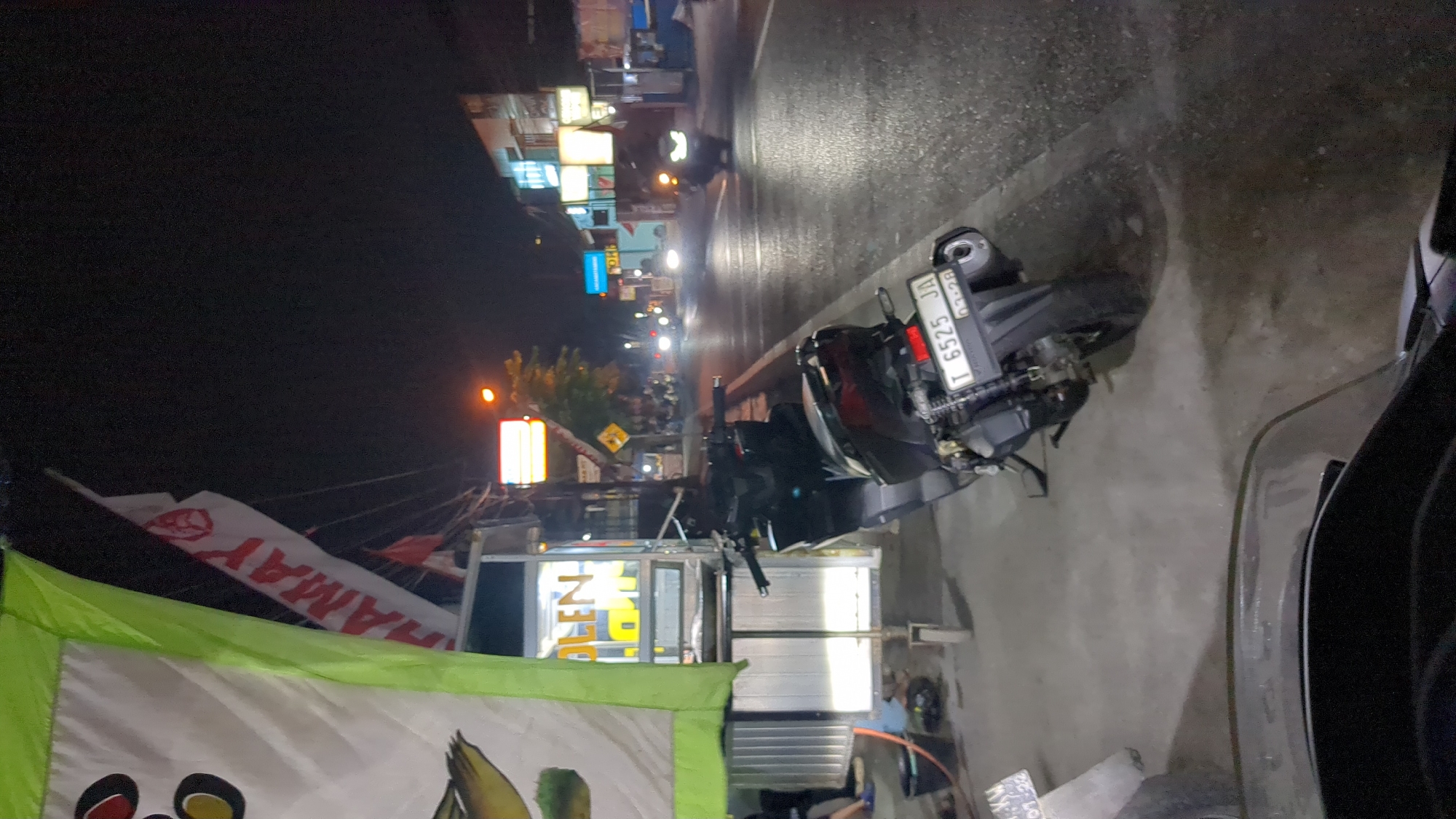 This is a nighttime photograph taken in Purwakarta, Indonesia, likely from a low angle, possibly from a moving vehicle. The scene depicts a street at night, illuminated by artificial lights from storefronts and streetlights. In the foreground, the handlebars and dashboard of a scooter or motorcycle are visible. To the left, a food cart with a green and white banner advertising "YAMAHA" is parked on the sidewalk. The cart has a metal frame and is lit from within, displaying some products. A black trash can is placed near the cart.
Further down the street, several motorcycles are parked along the sidewalk and on the road. One motorcycle in the mid-ground has a license plate that reads "T 6525 JA" with a date of "03-28". Other motorcycles are seen in motion, with their headlights on, suggesting traffic on the road. Storefronts with illuminated signs line the right side of the street, displaying a variety of colors and shapes, indicating a commercial area. The road surface appears wet, reflecting the lights and suggesting recent rain or a damp environment. The overall atmosphere is that of a bustling street scene at night in an urban setting.