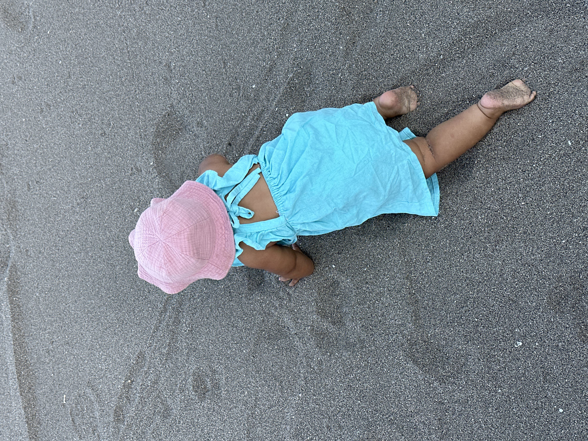 A baby girl wearing a pink sun hat and a turquoise dress is crawling on a dark sandy beach. Her legs and feet are dirty from the sand. The scene is likely located on a beach in North Kuta, Indonesia.