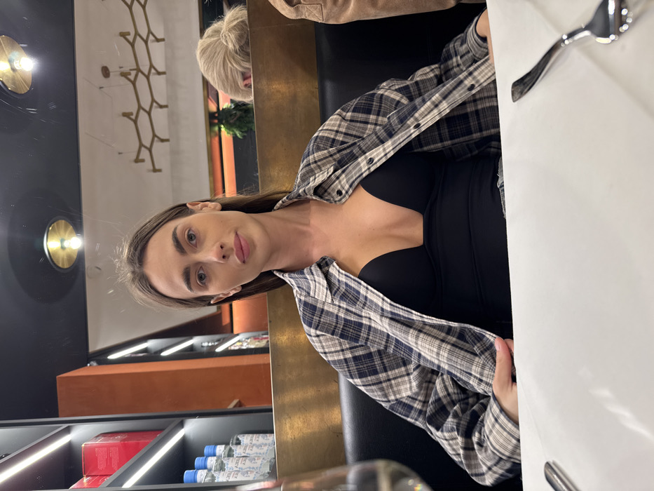 An indoor scene in a restaurant or bar in Verona, Italy, depicts a young adult female seated at a table, facing the camera directly. She has long, straight brown hair, light eyes, and neutral facial expression with slightly parted lips. She is wearing a deep-V black top beneath an unbuttoned plaid shirt in shades of blue, white, and brown.
The woman is seated in a dark booth with a golden-brown backrest. A white tablecloth covers the table in the foreground. A wine glass, partially visible on the left, contains a clear liquid. A silver fork rests on the right side of the tablecloth.
In the background to the left, a tall shelving unit with vertical LED strip lighting displays multiple bottles with blue caps and white labels, some appearing to be "LUCKY" brand, alongside red boxed items on an upper shelf. The background to the right features a large mirror reflecting two circular, gold-toned overhead light fixtures and a decorative, geometric chandelier. A portion of another individual's head, characterized by blonde hair tied in a bun, is visible in the reflection behind the primary subject. The ceiling is dark.