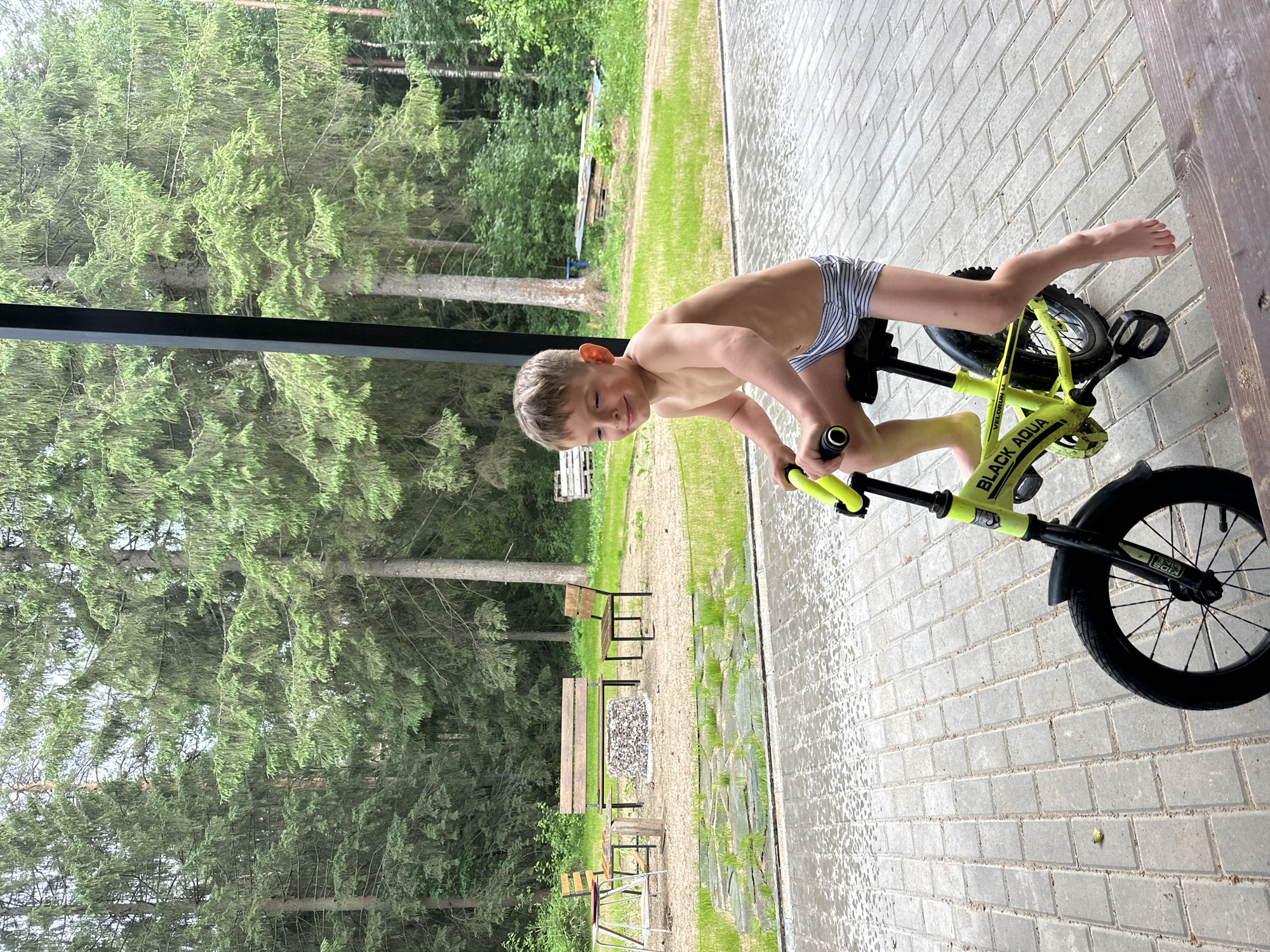 A young, shirtless boy with light brown hair, wearing striped shorts, is sitting on a bright yellow balance bike. He is on a paved outdoor area made of grey rectangular bricks, which appears wet. The boy is looking to his right with a slight smile, and his right hand is on the bike's handlebars. In the background, there are tall green pine trees, a grassy area, a gravel fire pit surrounded by benches and chairs, and some wooden structures. The scene is likely a backyard or recreational area in a wooded setting, possibly in or near the city of Perm (Прикамский refers to the region along the Kama River, which Perm is a part of), Russia.