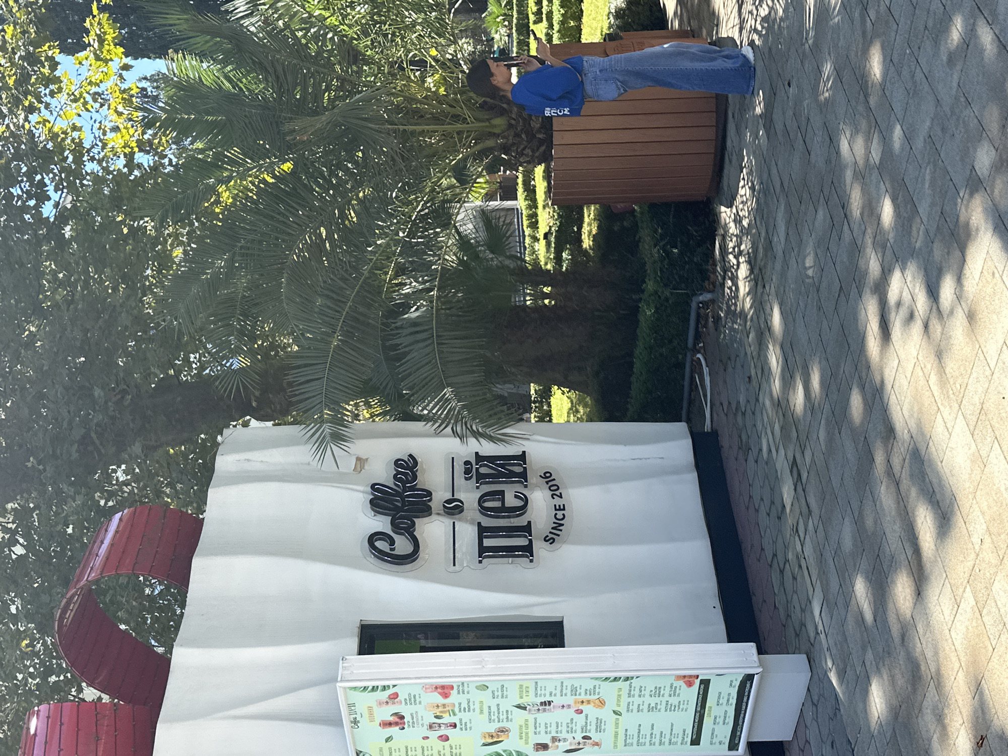 The image shows a "Coffee Пей" stand in Sochi, Russia, with a woman in a blue shirt and jeans standing next to a large planter. The stand is white with a red ribbon design on top, resembling a gift box. A menu is displayed on the left side of the stand. The background features palm trees and greenery, suggesting a park or outdoor seating area. The woman is holding a phone and appears to be looking at it. The ground is paved with cobblestones, and there are shadows cast by the trees and the stand.