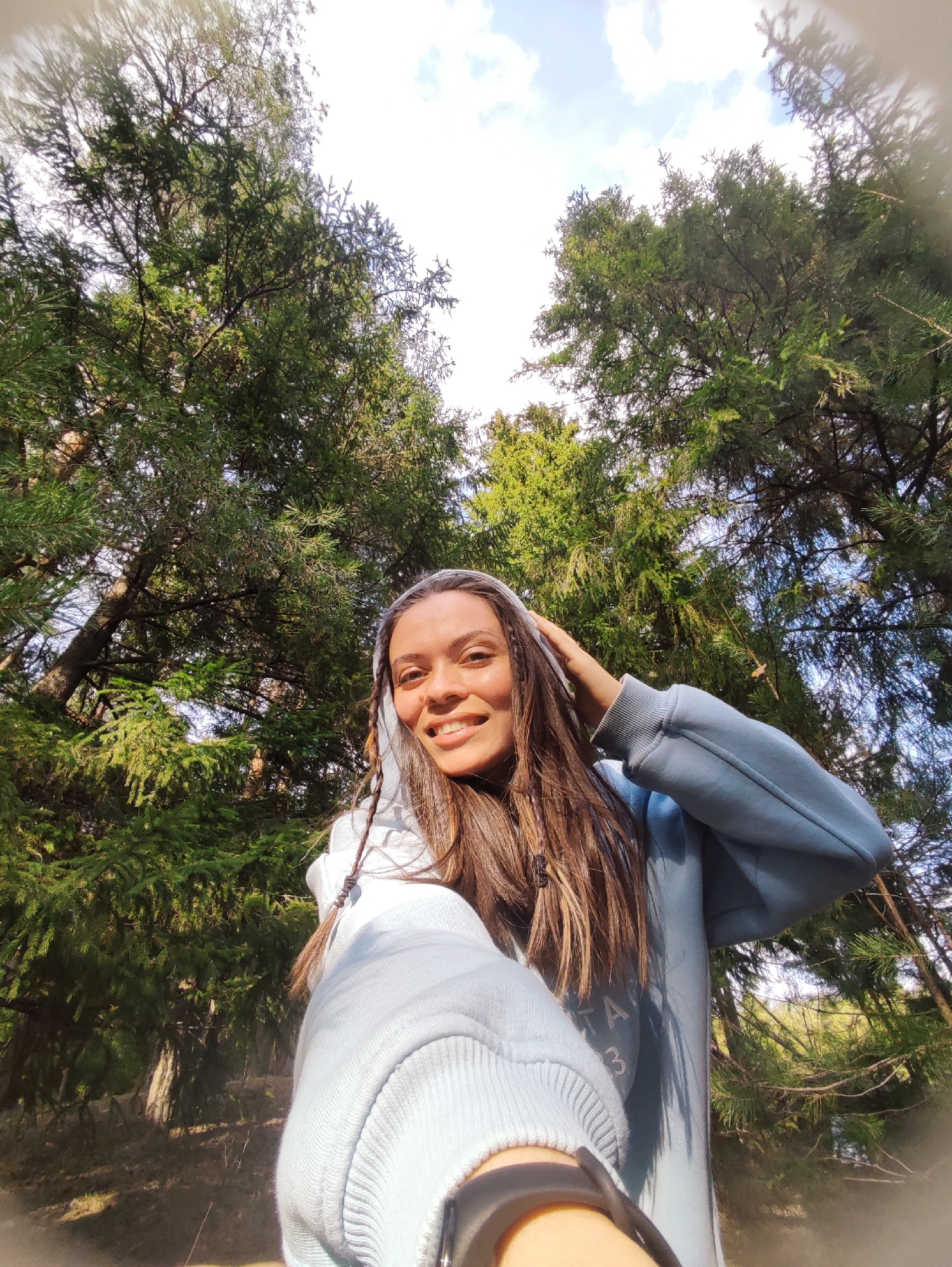 A smiling woman stands in a forest, likely taking a selfie. She is wearing a light blue hooded sweatshirt and has long brown hair with some braids. Her hand is raised to her head. Tall evergreen trees surround her, filling most of the frame. The sky is visible between the trees, showing some clouds. The location, Фоки, Russia, cannot be verified from the image.