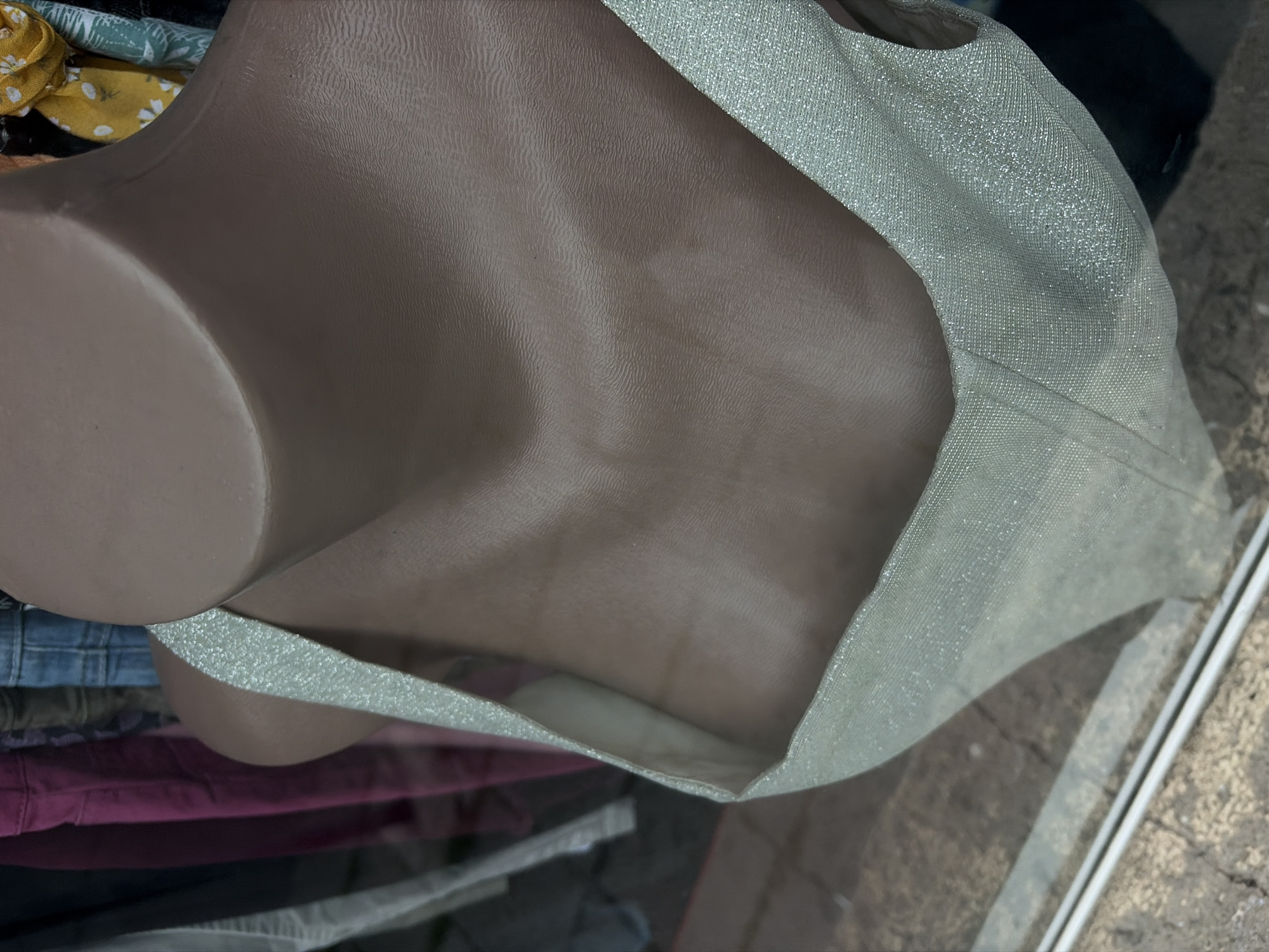 The image shows a mannequin torso wearing a glittery silver top, possibly displayed in a shop window. The bottom of the mannequin is partially obscured by a stack of clothing. The background includes a glimpse of a street scene with pavement, suggesting the location is likely in a retail area, possibly Burgas, Bulgaria.