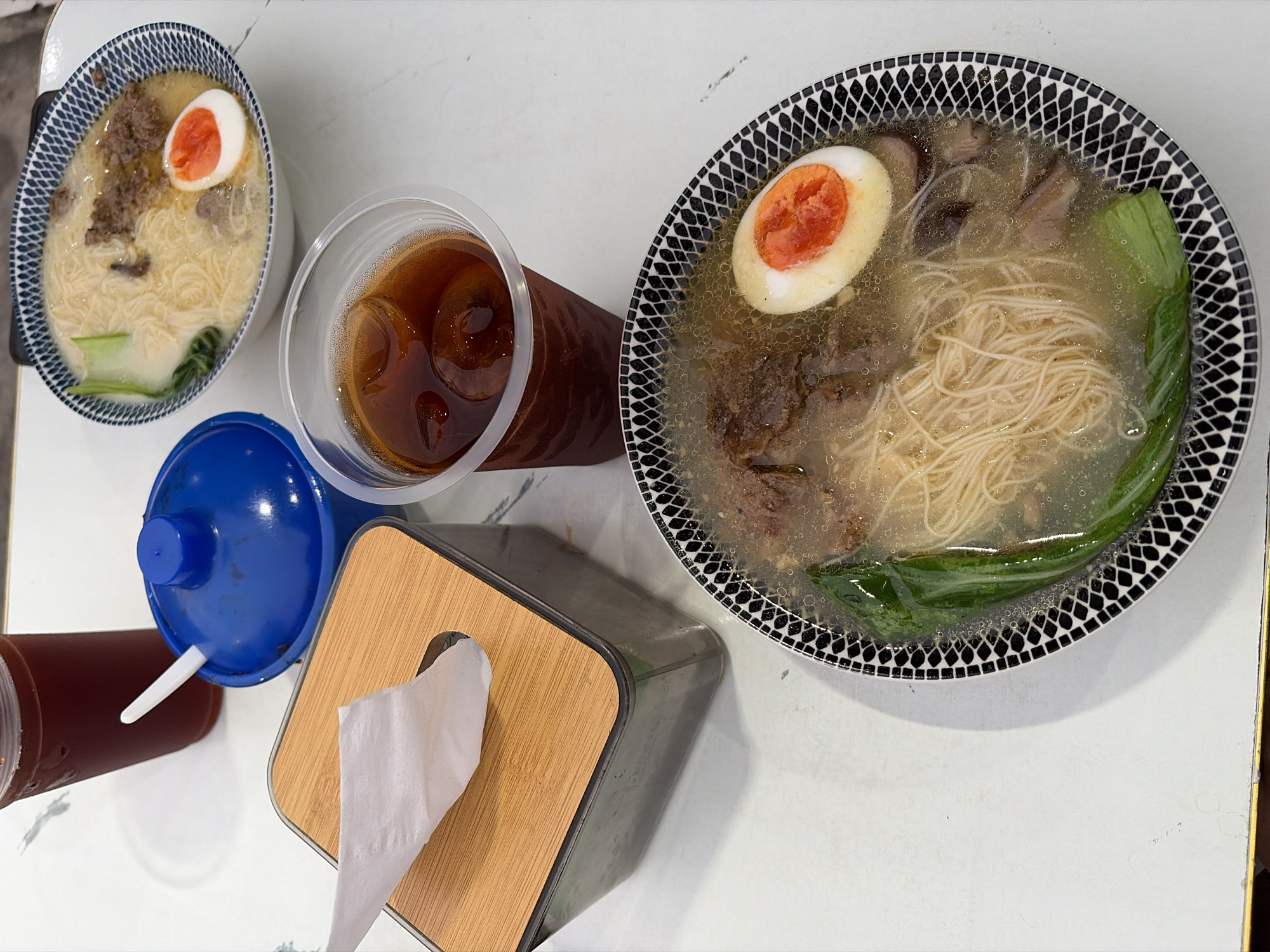 The image shows a table setting with two bowls of ramen, two drinks, a tissue box, and a blue container with a spoon. The ramen bowls appear to contain noodles, broth, meat, and a hard-boiled egg. One bowl has a decorative black and white pattern around the rim, while the other has a more intricate blue and white pattern. The drinks are in clear plastic cups, one filled with a dark liquid and ice, and another taller glass with a reddish-brown liquid. The tissue box is made of wood and plastic, with a white tissue protruding. The blue container is round and has a lid. The scene is set on a white, marble-like surface. The overall impression is a casual meal, possibly at a restaurant. Based on common Semarang cuisine, the ramen dishes are likely representative of local Indonesian adaptations of this popular dish, often found in eateries serving a variety of Asian comfort foods in cities like Semarang.