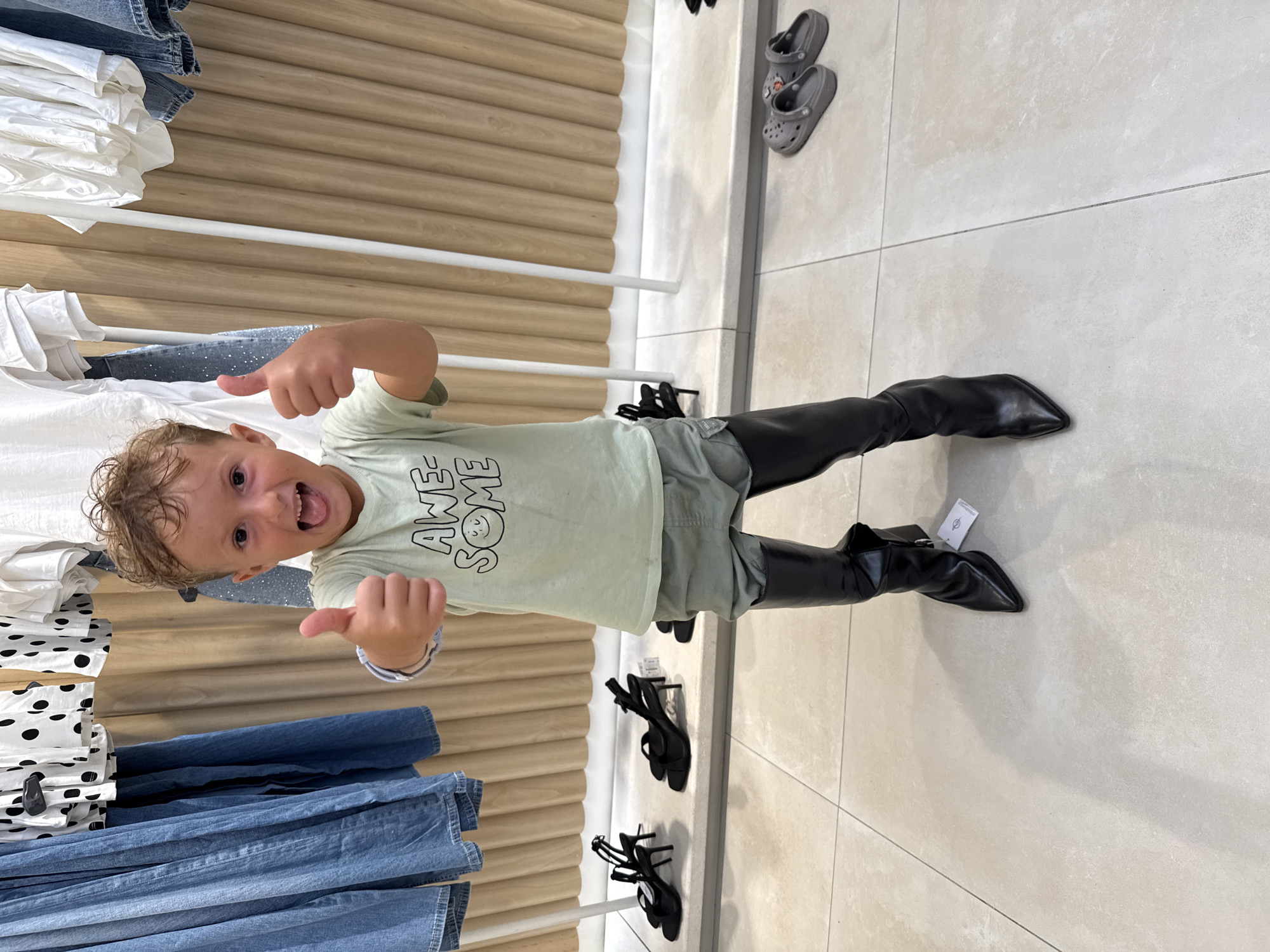 A young boy, appearing to be around 4-5 years old, stands in what appears to be a retail store. He is wearing a light green t-shirt with the words "AWESOME SMILE" printed on it, and light grey shorts. He is also wearing a pair of oversized, black, pointed-toe knee-high boots. The boy is looking at the camera with a wide smile and his tongue sticking out slightly. He has his thumbs raised in a positive gesture. In the background, clothing racks with various garments, including denim jeans and white tops, are visible. On a lower shelf, a pair of black high-heeled sandals are displayed. Further in the background, on the tiled floor, a pair of grey Crocs are visible. The floor is tiled with large, light-colored square tiles. The overall environment suggests a clothing store, likely in a shopping area. The location of Kuta, Indonesia, is not discernible from the image itself, but is provided as context.
