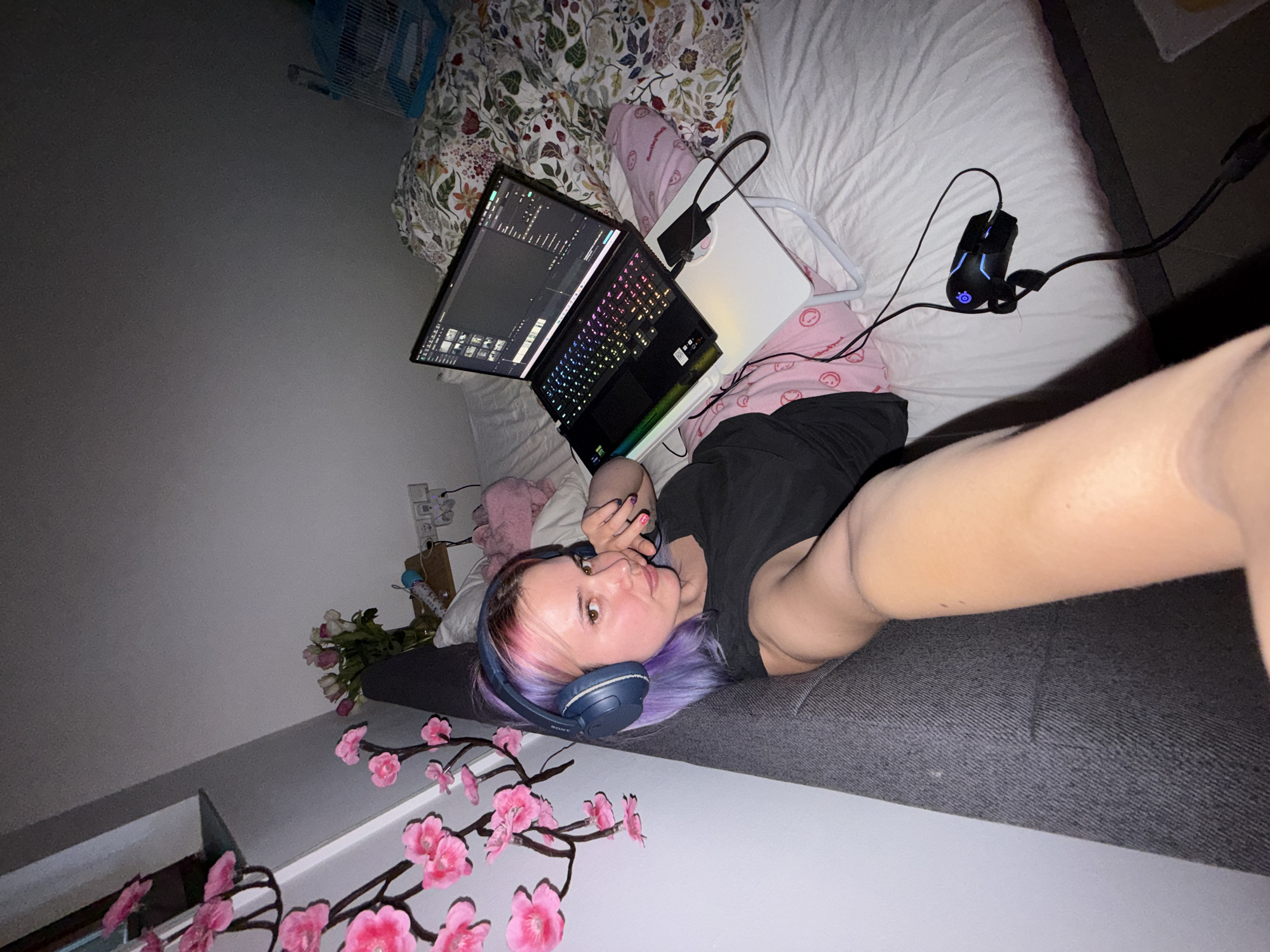A female with light skin, purple and pink-rooted hair, and blue over-ear headphones is depicted lying on her left side on a bed, taking a selfie. She wears a dark t-shirt and pink pajama pants with white smiley face patterns. Her right arm is extended towards the camera, while her left hand rests near her chin. She is propped against a dark grey upholstered headrest or pillow.
A black gaming laptop with an RGB backlit keyboard is open on a white foldable lap desk, displaying video editing software. A black external drive is connected to the laptop on the desk. A wired black gaming mouse with blue LED accents (SteelSeries brand) lies on the white bedsheet to the right of the lap desk. The bed is covered with a white fitted sheet and a floral duvet featuring red, green, and white patterns on a dark background.
To the left, against a light grey wall, is a decorative branch adorned with pink artificial cherry blossoms. Behind the female's head, on a wooden wall shelf, a vase contains a bouquet of white and pink flowers. An electrical outlet with multiple plugs is visible below the shelf. In the background, on the right side of the bed, a light blue and white birdcage is present. The room is dimly lit, primarily by the laptop screen and keyboard. This scene takes place in a bedroom or living space within the City of Zagreb, Croatia.