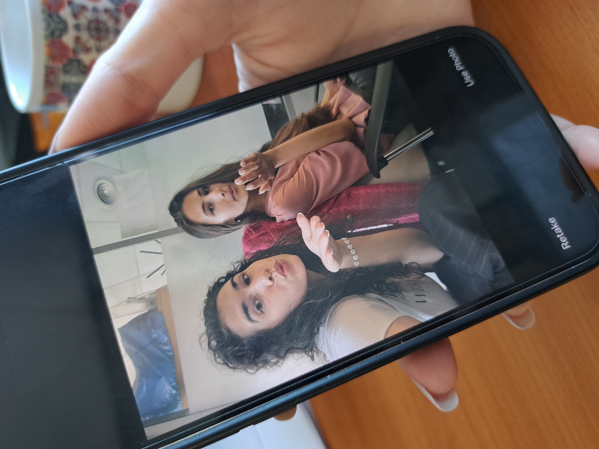 A hand holds a black smartphone, presenting a horizontal view of its screen. The screen displays a photo of two young women, both looking towards the camera. The woman in the foreground has dark, curly hair and is making a playful "kissy" face, with one hand raised, wearing a white t-shirt. She also wears a light-colored beaded bracelet. Behind her, another woman with long, straight dark hair looks directly at the camera with a slight smile. She is dressed in a pink long-sleeved top and a reddish, textured jacket. The photo appears to be taken indoors, possibly a mirror selfie or a photo taken in a room, with reflections of a ceiling vent, a dark chair, and a blue object visible within the screen's edges.
On the smartphone interface, the text "Use Photo" is visible in white at the top right corner, and "Retake" is partially visible at the bottom right, suggesting the user is reviewing a recently captured image. The hand holding the phone has light-colored nails. In the blurred background outside the phone, a portion of a colorful, patterned mug is visible, sitting on a wooden surface. The overall scene captures the act of reviewing a casual personal photograph.