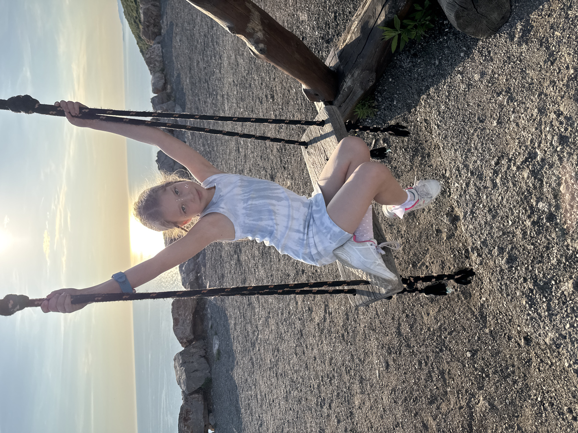 A young girl is captured mid-swing on a rustic wooden swing set during the golden hour in Kovačko Polje, Montenegro. She is the central subject, posing with her arms outstretched, holding onto the thick, dark ropes. She wears a light blue tie-dye tank top and shorts, white sneakers with pink accents, and has glitter adornments on her face. Her legs are bent, and she looks directly at the camera with a gentle smile.
The swing set is constructed from rough, natural wooden logs. The ground beneath is covered in small grey gravel and stones. In the background, the sun is low on the horizon, casting a warm, golden glow across a vast body of water, likely the Adriatic Sea, and a clear sky with soft clouds. Distant land or mountains are faintly visible across the water. The warm light creates a serene and cheerful atmosphere, highlighting the joy of the activity at sunset. There is no visible text in the image.
