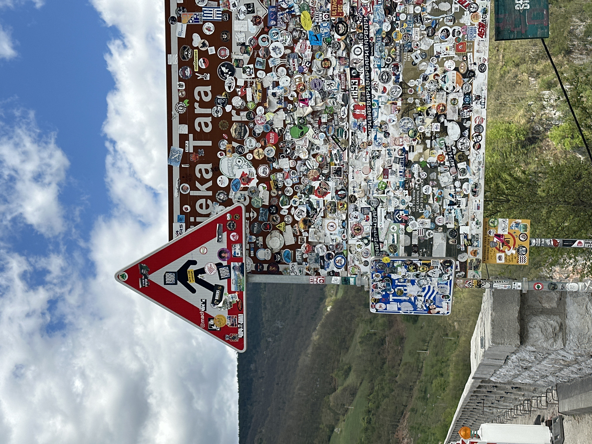 A roadside view in Đurđevića Tara, Montenegro featuring a signpost covered in numerous stickers. The signpost is comprised of several signs, including a red and white pedestrian crossing sign. Above it is a sign reading "Neka Tara" in white letters on a brown background. Further signs are covered in stickers from various locations and groups. The background consists of a mountain landscape, trees, a cloudy sky, and a section of road infrastructure.