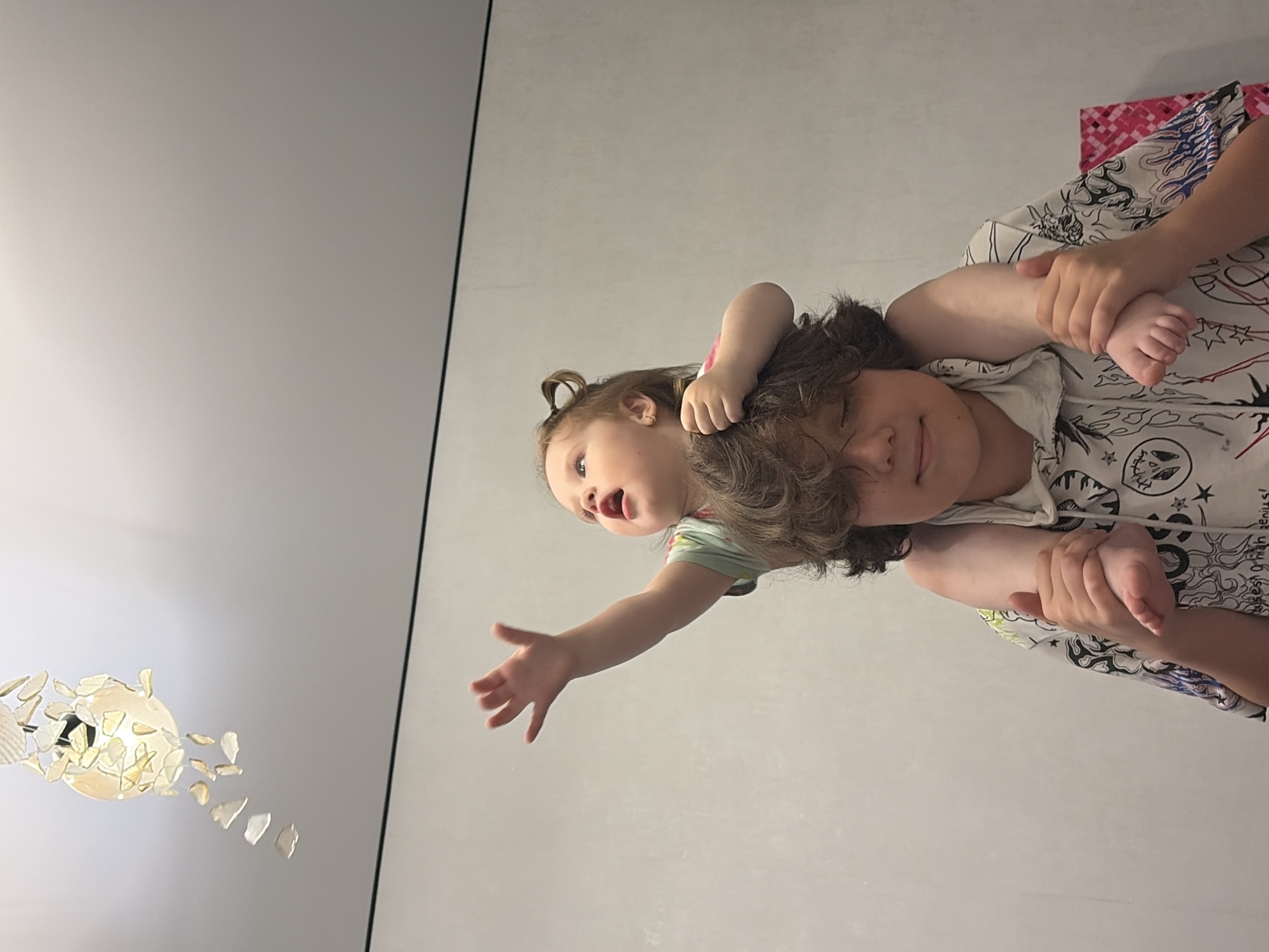 A young child, likely a toddler, is perched on the shoulders of an older child, who is looking downwards and to the right. The toddler is reaching upwards with their right arm, their hand open, towards a decorative light fixture hanging from the ceiling. The background consists of a plain, light-colored wall with a thin black line near the top. The overall lighting suggests an indoor setting. The image does not contain any information that would indicate a specific location in Chaikovsky, Russia.