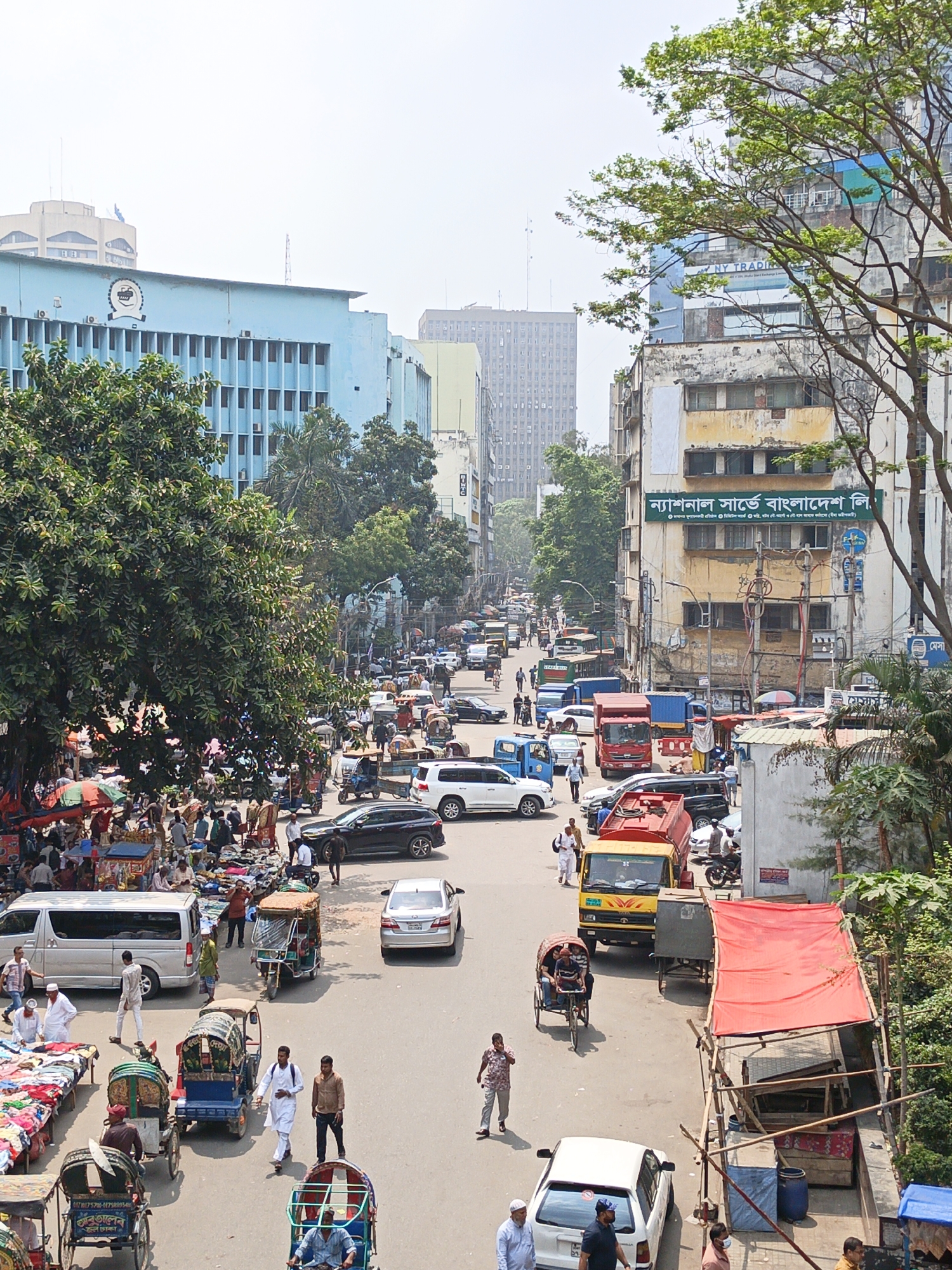 The image depicts a bustling street scene in Dhaka, Bangladesh, viewed from an elevated perspective. The street is filled with a mix of vehicles, including cars, buses, trucks, and cycle rickshaws, also known as pedicabs. Pedestrians walk along the street and sidewalks, with some gathered around what appears to be a market area on the left side of the frame. Buildings of varying heights line the street, including a light blue building with a clock on its facade and a sign indicating "BIWTC". Another building displays signage in Bengali script, potentially related to a surveying company: "ন্যাশনাল সার্ভে বাংলাদেশ লি.". The sky is overcast, and the scene suggests a typical busy day in a dense urban environment.