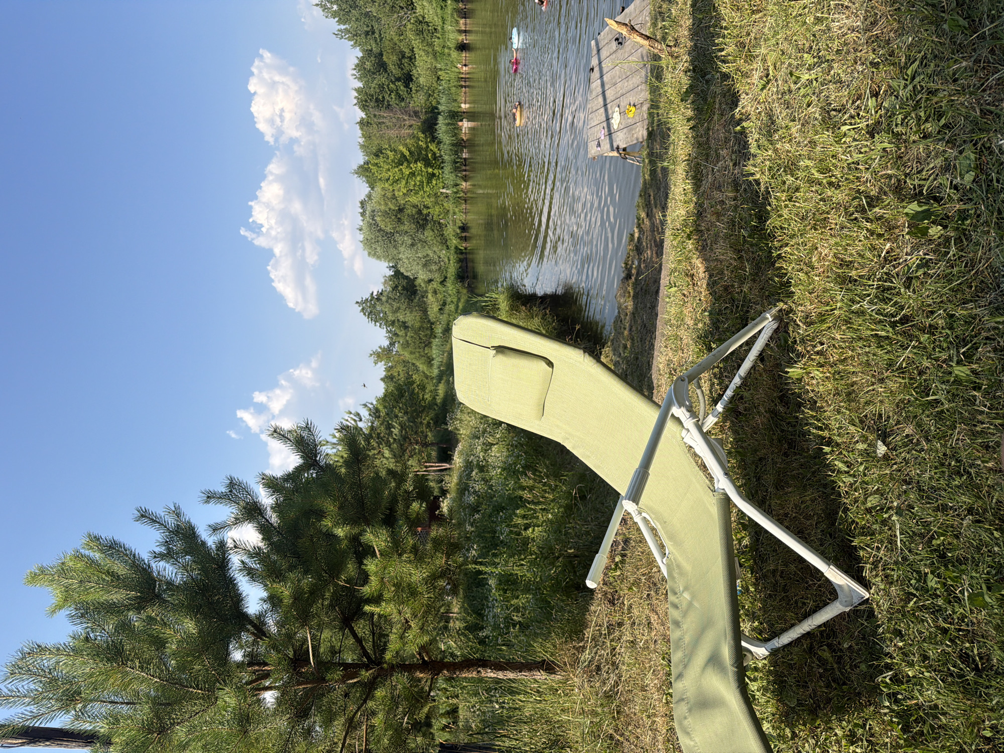 This image depicts a serene outdoor scene likely in Kamianka-Buzka, Ukraine, featuring a green lounge chair placed on a grassy area next to a calm body of water. The water is bordered by lush green trees and reeds, with a wooden pier extending into the water. Several inflatable tubes or swimming aids are visible in the water, suggesting recreational activity. The sky is a clear blue with scattered white clouds. The overall impression is one of a tranquil summer day in a natural setting.