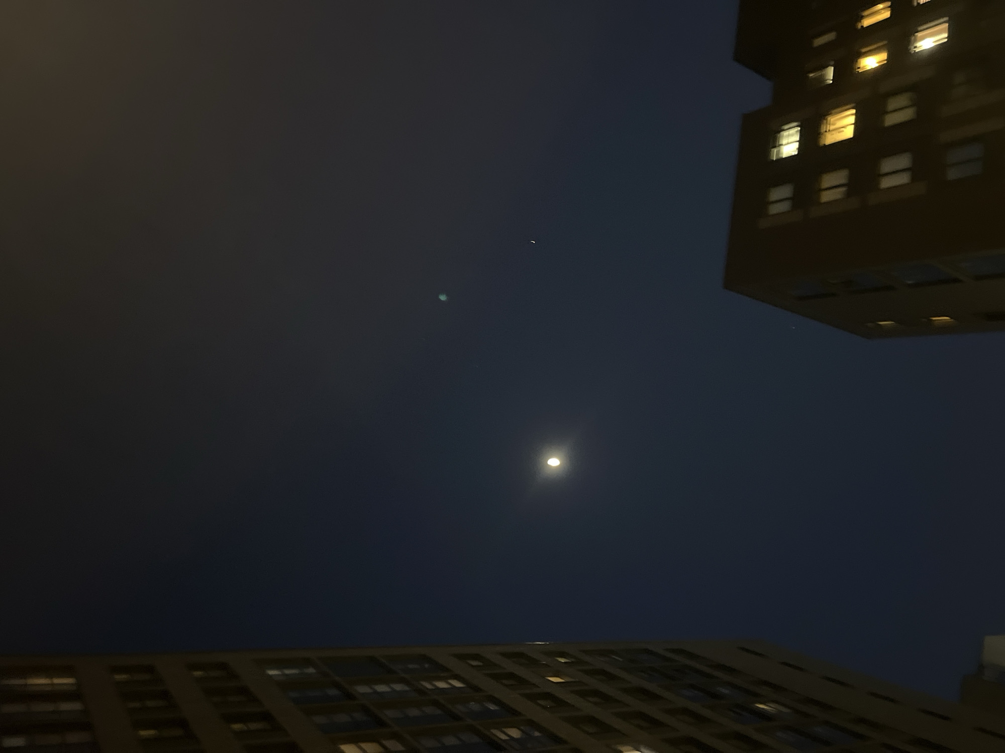 The image presents a nighttime scene in Saint Petersburg, Russia, viewed from a low angle between two buildings. The dominant feature is the dark, cloudless sky, which contains a prominent, bright, circular moon. Above and to the right of the moon, two fainter light anomalies are visible: a diffuse greenish circular glow and a smaller, distinct white crescent shape, consistent with lens flare artifacts.
To the left, a multi-story building with a light-toned facade extends vertically, partially cropped at the top and bottom. Its surface is punctuated by numerous rectangular windows, with several displaying warm yellow interior illumination. In the lower right quadrant, another multi-story building with a darker-toned facade is visible, also featuring multiple rectangular windows from which warm yellow light emanates. No people, vehicles, or ground-level specificities are identifiable in the frame.