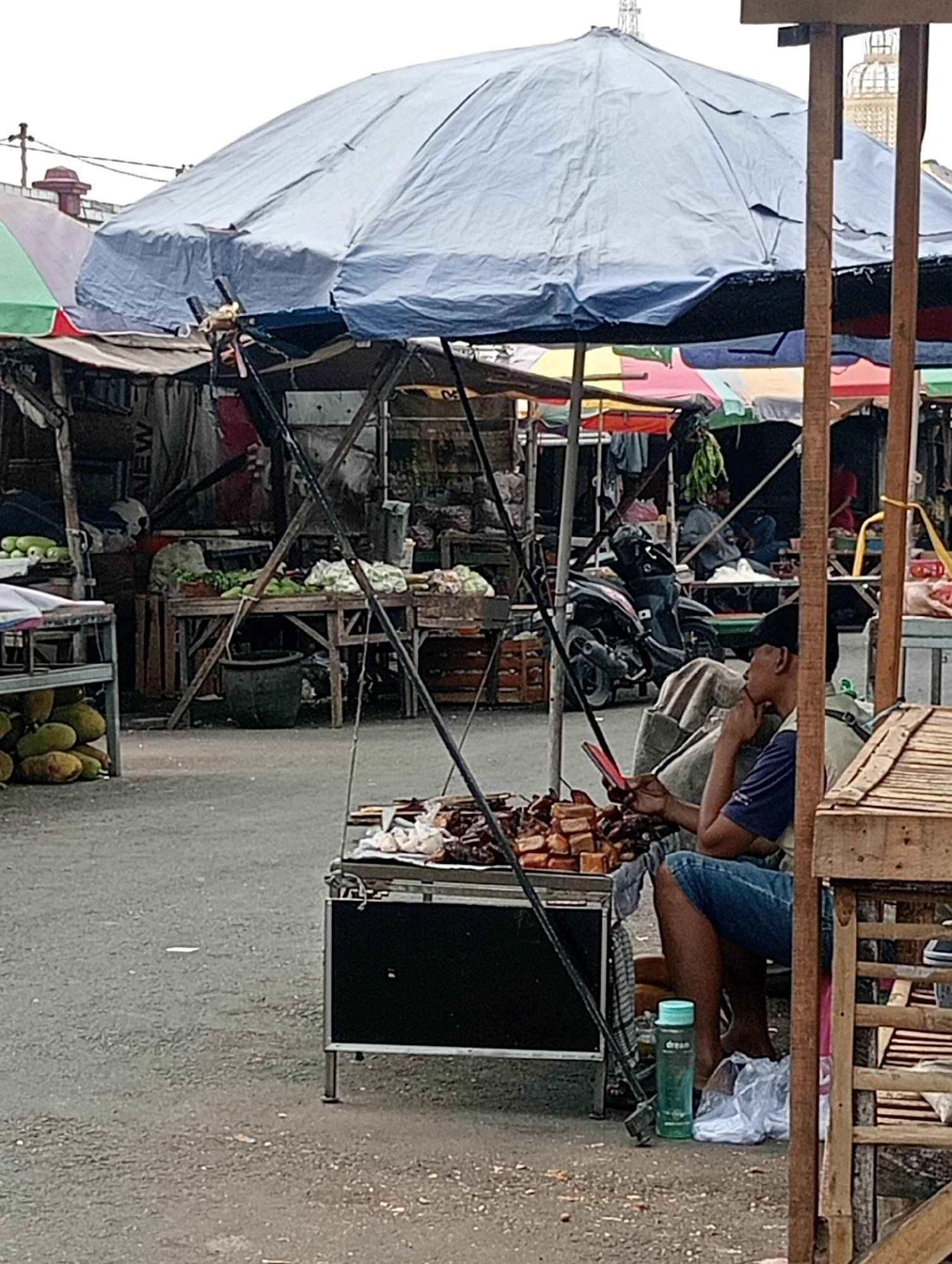 A street scene in the city of Jagalan, Indonesia, depicts an outdoor market with several stalls. A large, faded blue tarp is stretched over a central stall, supported by metal poles. Various colorful awnings are visible behind it, suggesting multiple vendors. In the foreground, a man wearing a black cap, blue shorts, and a blue and white shirt sits on a stool, holding a red mobile phone. He appears to be looking at the phone and resting his chin on his hand. In front of him, on a black wheeled cart, is a display of food items, possibly grilled meats or skewers, arranged in neat rows. A green water bottle is placed on the ground next to the cart, beside a plastic bag. To the left of the central stall, another stall displays produce, including jackfruit and what appear to be corn. A motorcycle is parked behind the central stall. The ground is paved with asphalt. The overall impression is of a bustling market atmosphere.