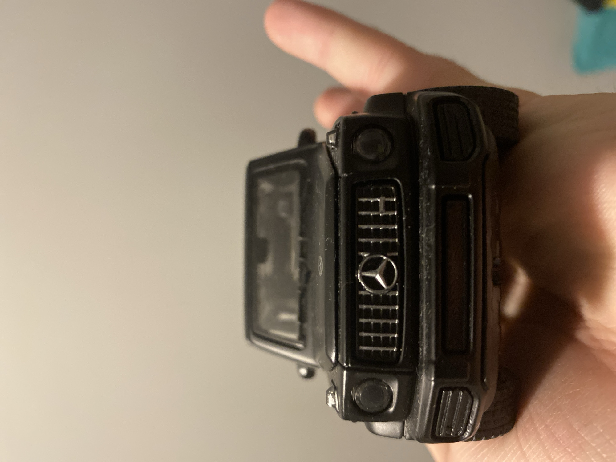 A black Mercedes-Benz G-Class toy car is held in a person's hand. The car is viewed from the front, with its grille and headlights clearly visible. The background is a plain, light-colored wall. There is no information in the image that places it in the city of Łódź, Poland.
