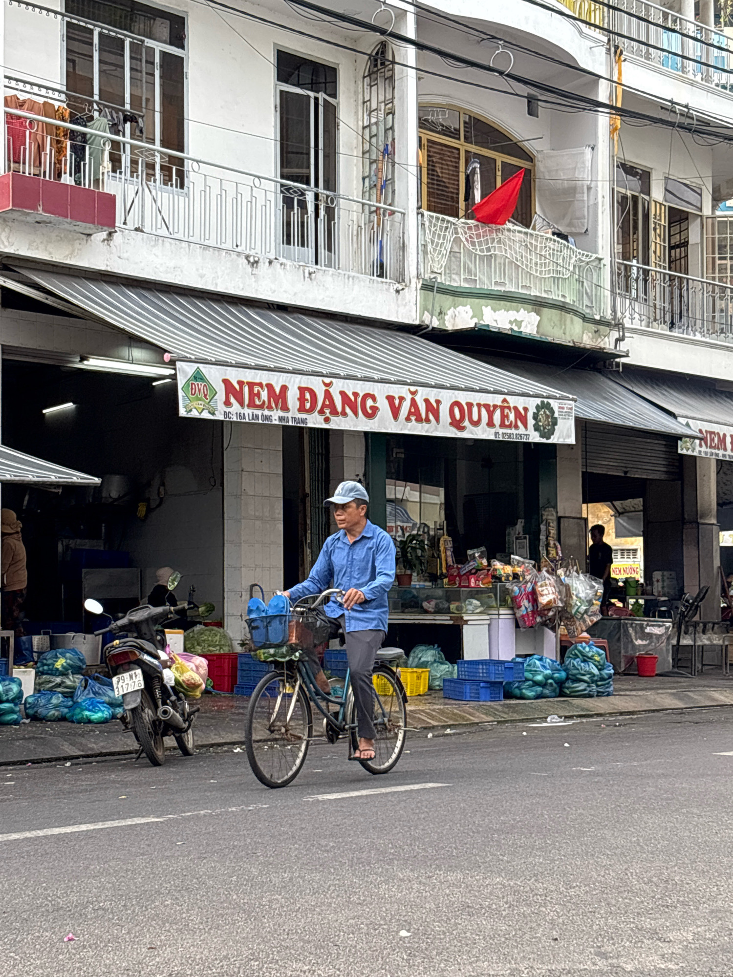 In Nha Trang, Vietnam, a man wearing a blue shirt and a cap rides a bicycle down a street lined with businesses. The man is positioned in the right half of the frame, pedaling his bicycle forward. His bicycle has a basket in the front, containing two blue containers. Behind the cyclist, a storefront displays various goods, including packaged items and what appear to be fresh produce. A motorcycle is parked to the left of the storefront. The street surface is asphalt, with a white line visible. The buildings in the background are multi-story structures with balconies and windows, suggesting an urban setting. The overall scene depicts a typical street view in a Vietnamese city, with commercial activity and everyday life unfolding.